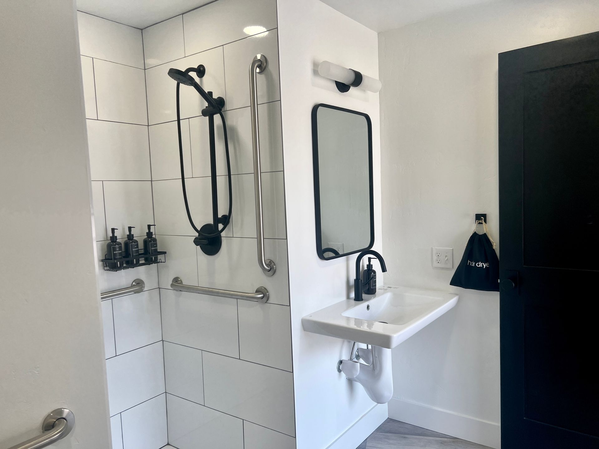 Accessible bathroom with shower and sink. White tile, black fixtures, and grab bars.