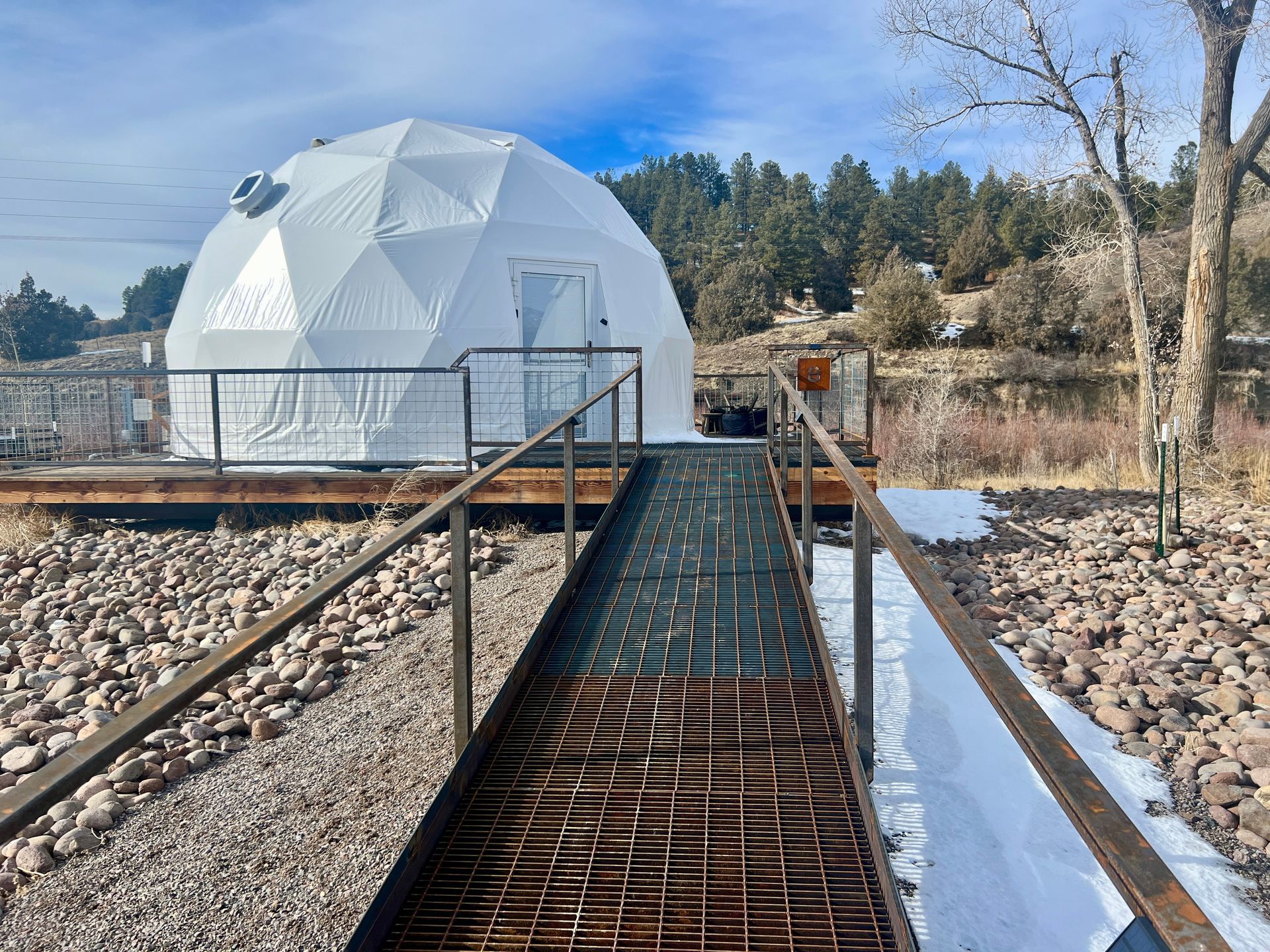 White geodesic dome structure, accessed by a metal ramp, in a natural outdoor setting with trees and snow.