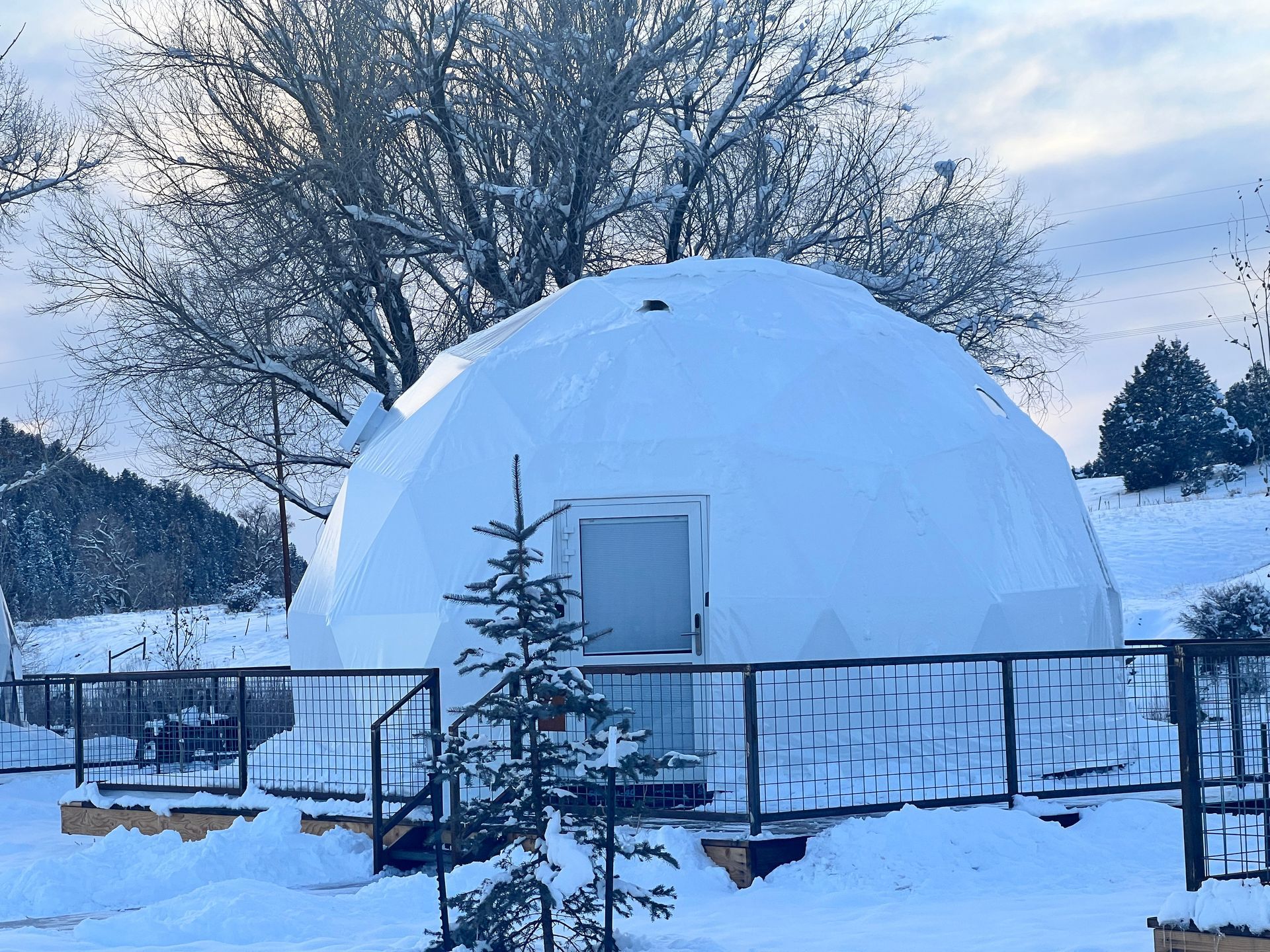 Snow-covered white geodesic dome structure with a door, in a snowy outdoor setting.