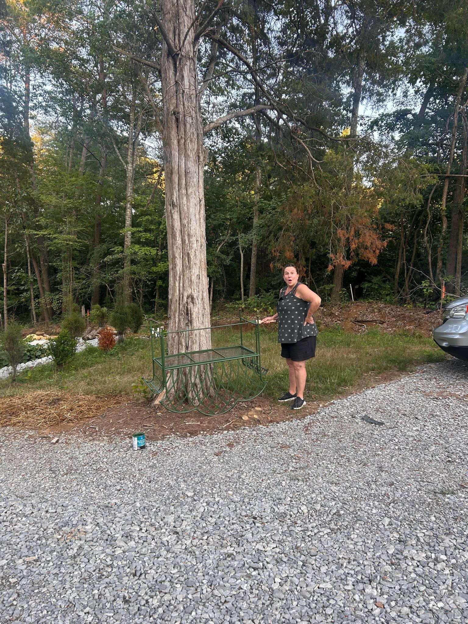 A woman is standing in a gravel driveway next to a tree.