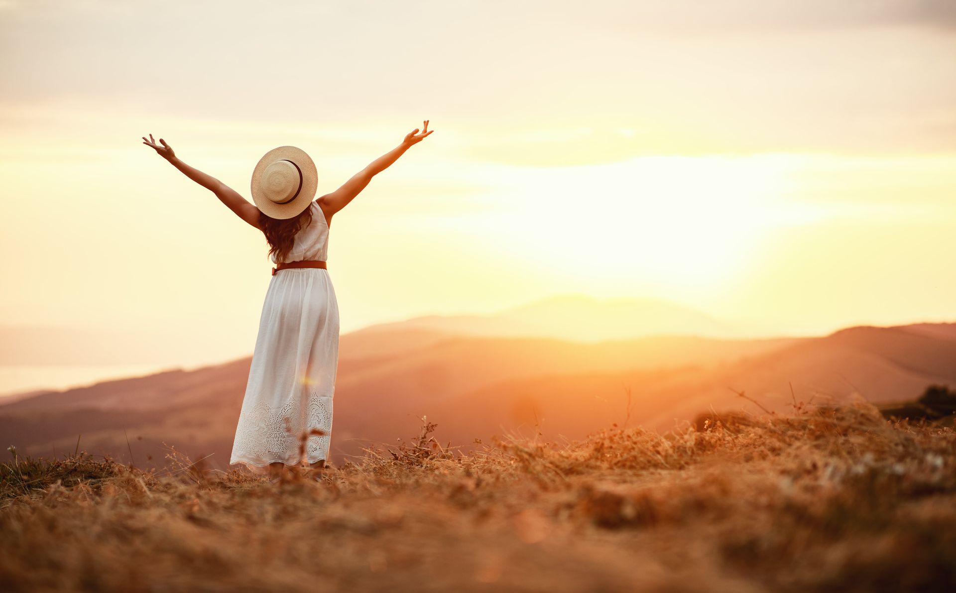 Uma mulher de vestido branco e chapéu está parada em um campo com os braços estendidos ao pôr do sol.