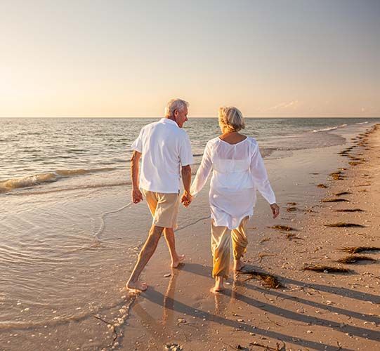 Elderly couple holding hands, walking on a beach at sunset, water and sand.