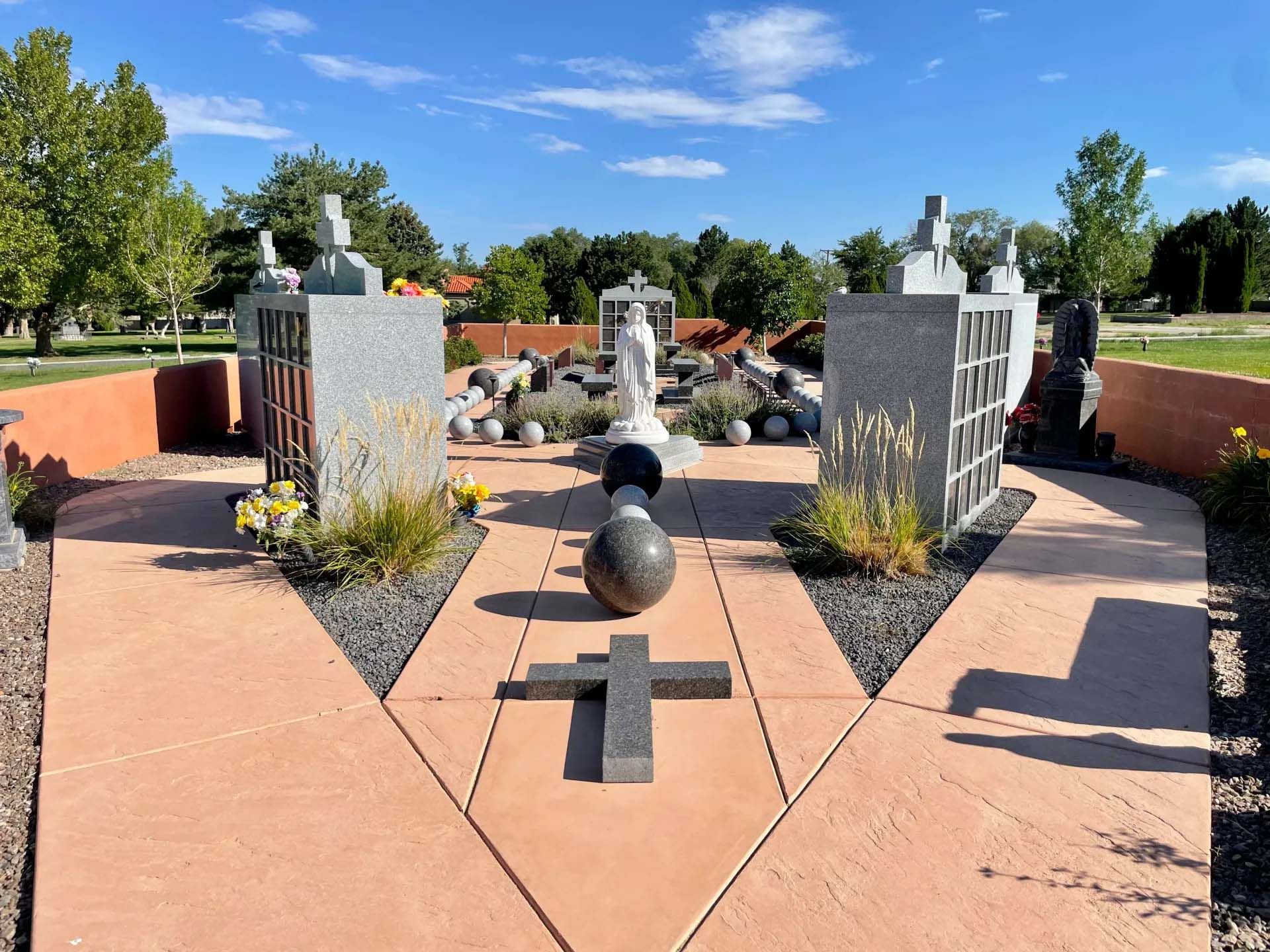 Cemetery with stone memorials, crosses, and statues. Paths lead to structures, under a blue sky.