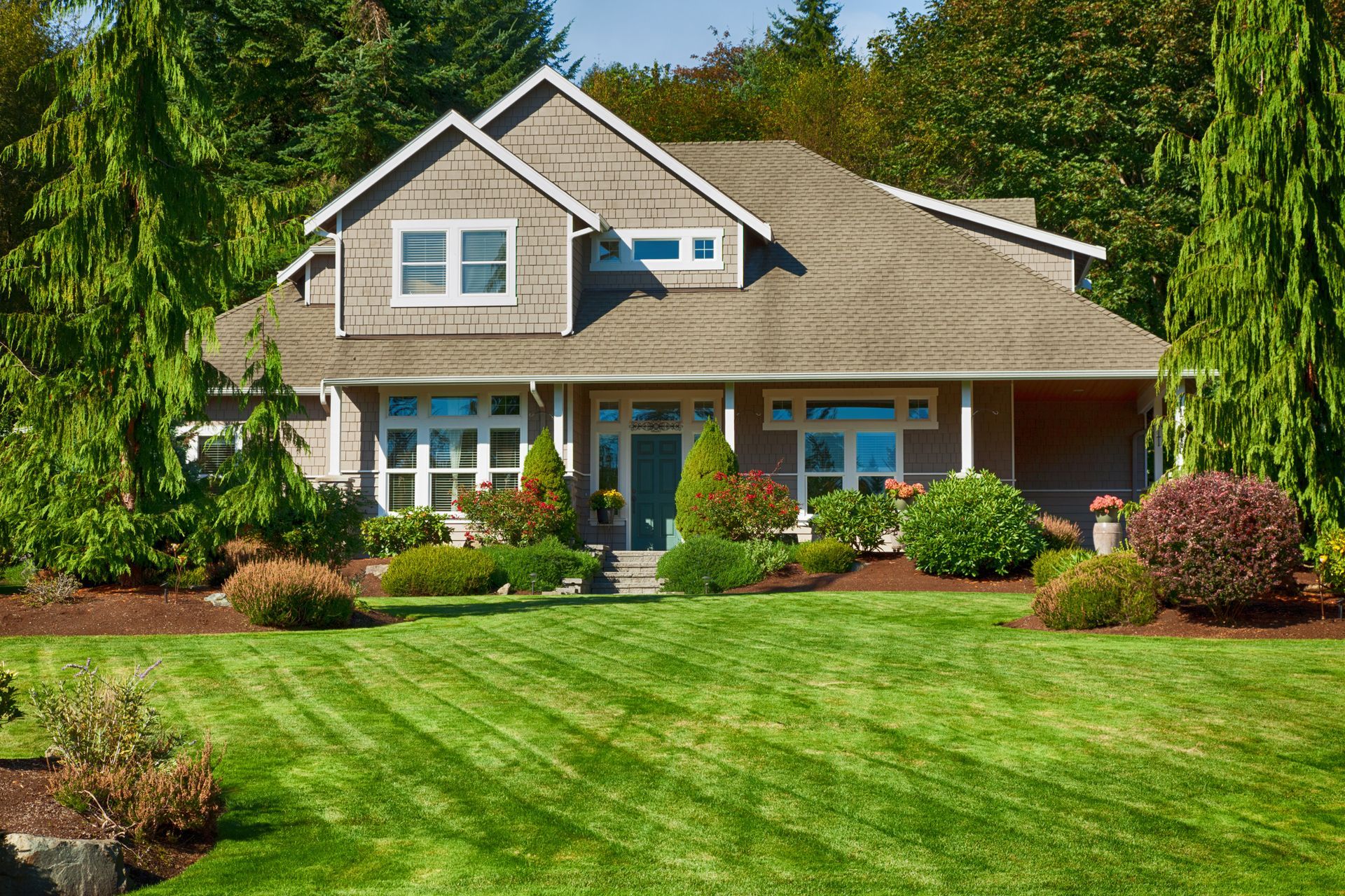 House with brown shingles, green lawn, landscaping, and a blue front door.