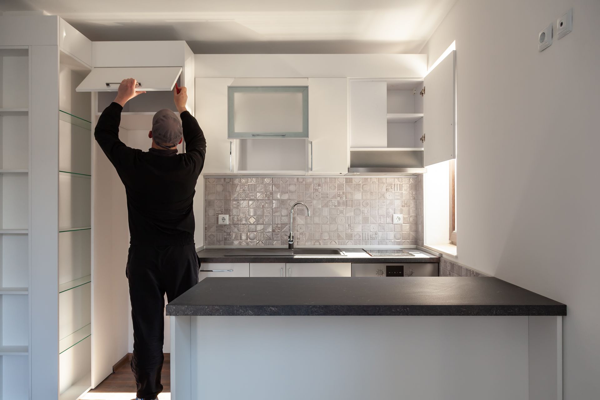 Person opening a white cabinet in a small, modern kitchen with a gray countertop and backsplash.