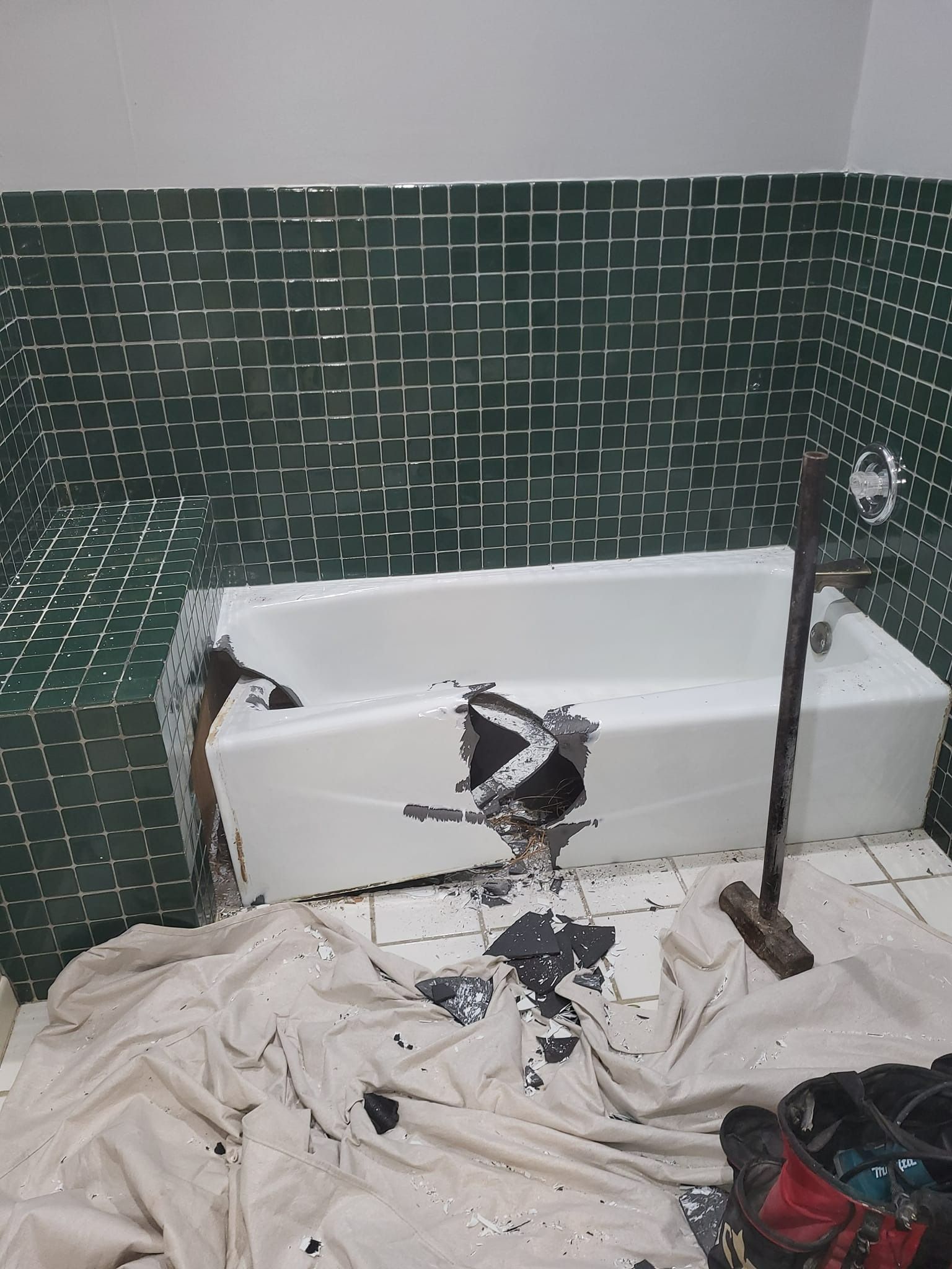 A damaged white bathtub with debris on the floor, surrounded by dark green tiled walls.