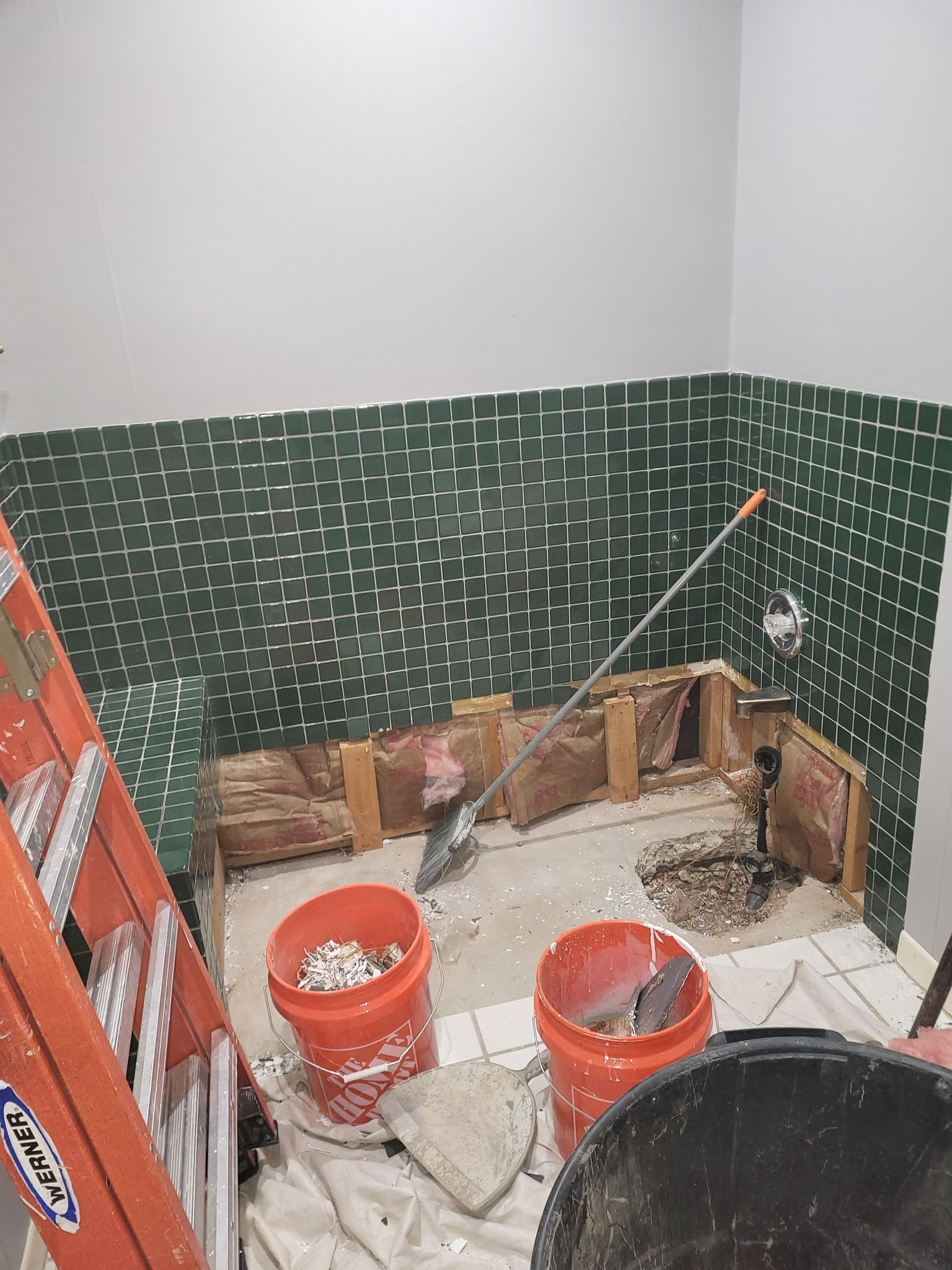 Bathroom renovation in progress. Green tiled wall, exposed studs, debris, buckets, ladder, and tools.