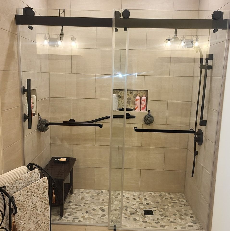 Shower with glass doors, stone tile floor and walls, black fixtures, and a small bench.
