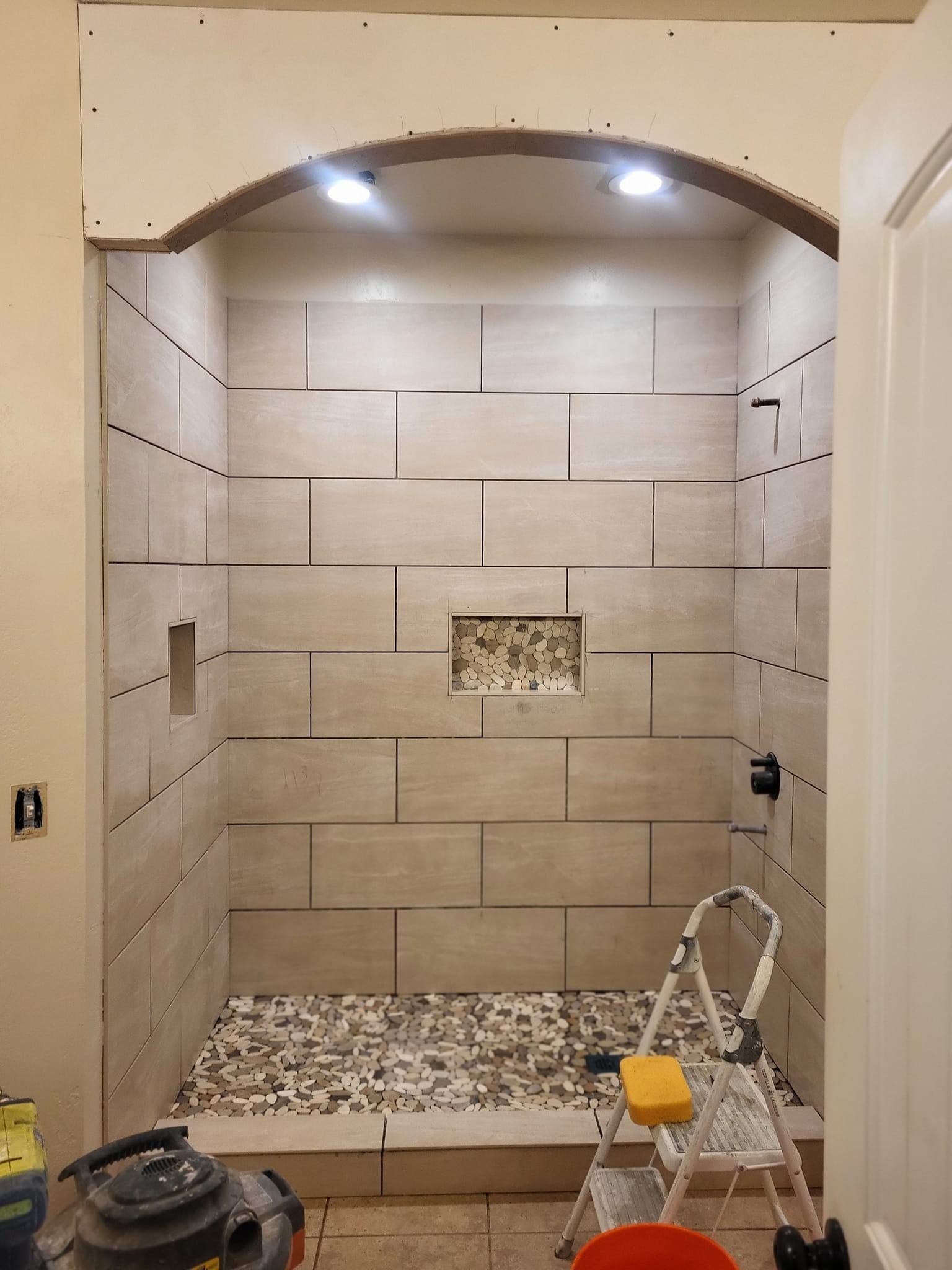Shower stall with stone tile, pebble floor, recessed shelf, two lights, step ladder.