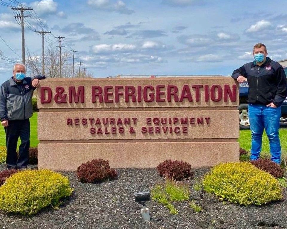 two men wearing masks stand in front of a sign for D&M Refrigeration