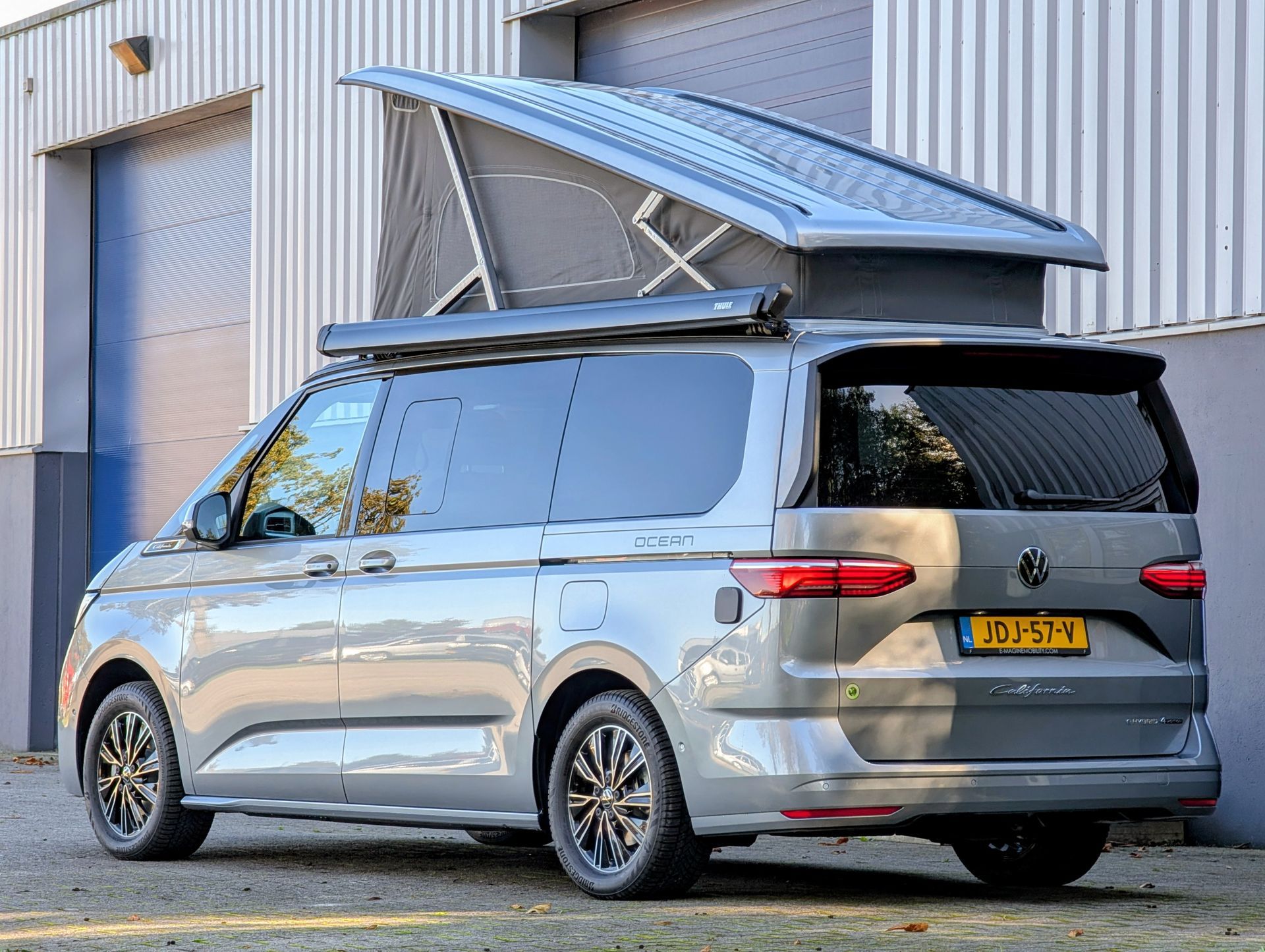 Volkswagen California Ocean eHybrid in de kleur Mono Silver, achteraanzicht met hefdak omhoog voor het gebouw van e-magine Mobility in Emmen