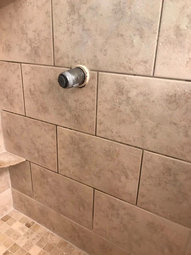Shower wall with beige rectangular tiles; a metal pipe protrudes through the tiles.