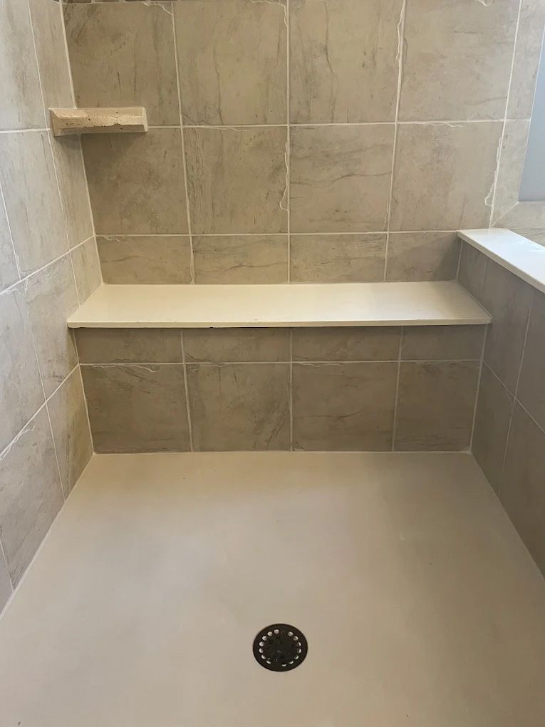 Shower with beige tiles, bench, and floor, a small shelf, and a drain.