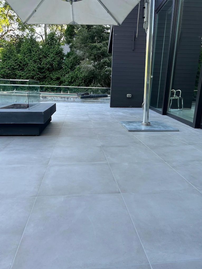 Grey tiled patio with a modern fire pit and dark building, umbrella to the side.