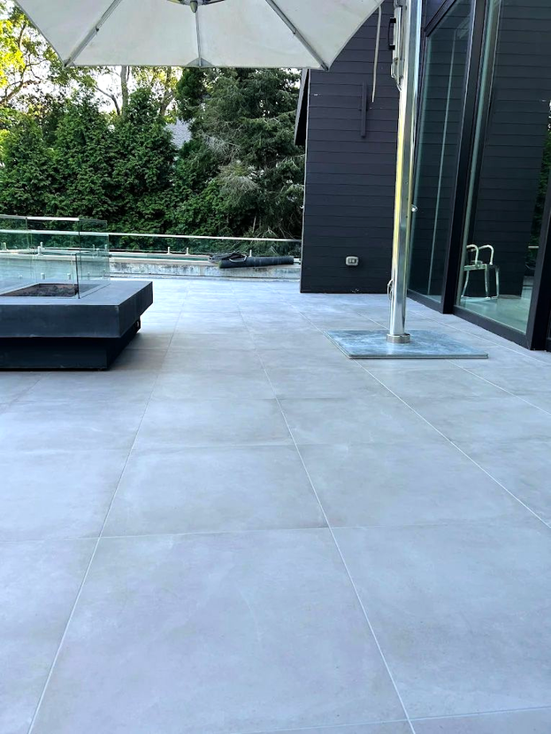 Gray tiled patio with a fire pit, umbrella, and modern black house with glass windows.