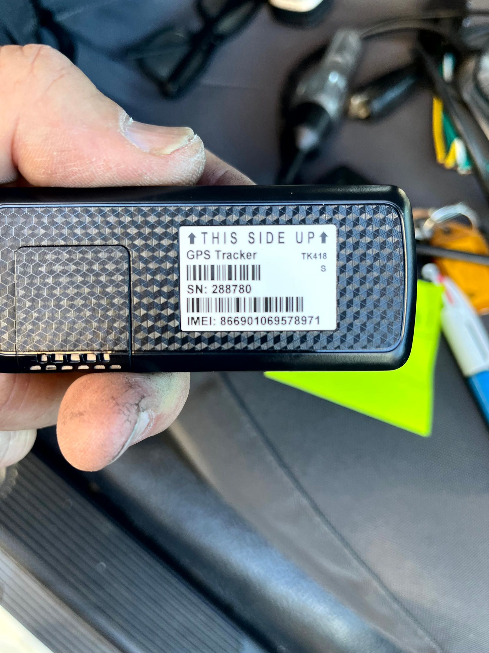 Hand holding a black GPS tracker with a label showing text and a barcode, on a car seat.