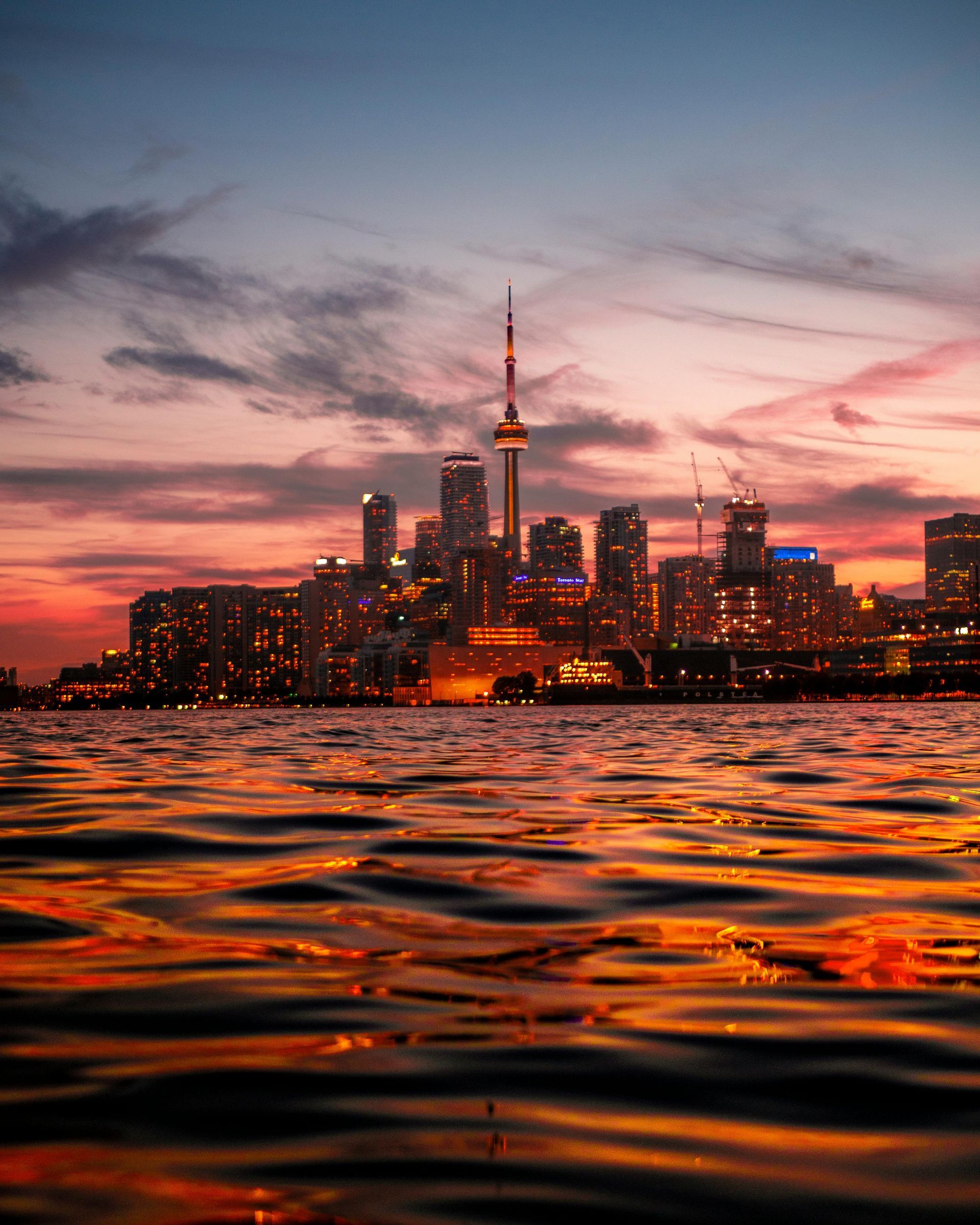 Toronto skyline at sunset, with CN Tower prominent, reflecting in water with orange and purple hues.