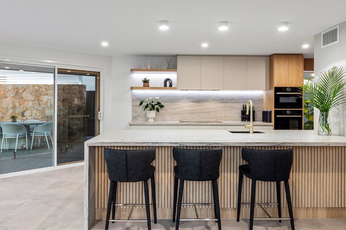 A Modern Kitchen With a Breakfast Bar, Three Black Stools, and a View to an Outdoor Patio Through Sliding Glass Doors — Truline Cabinets In Tuchekoi, QLD