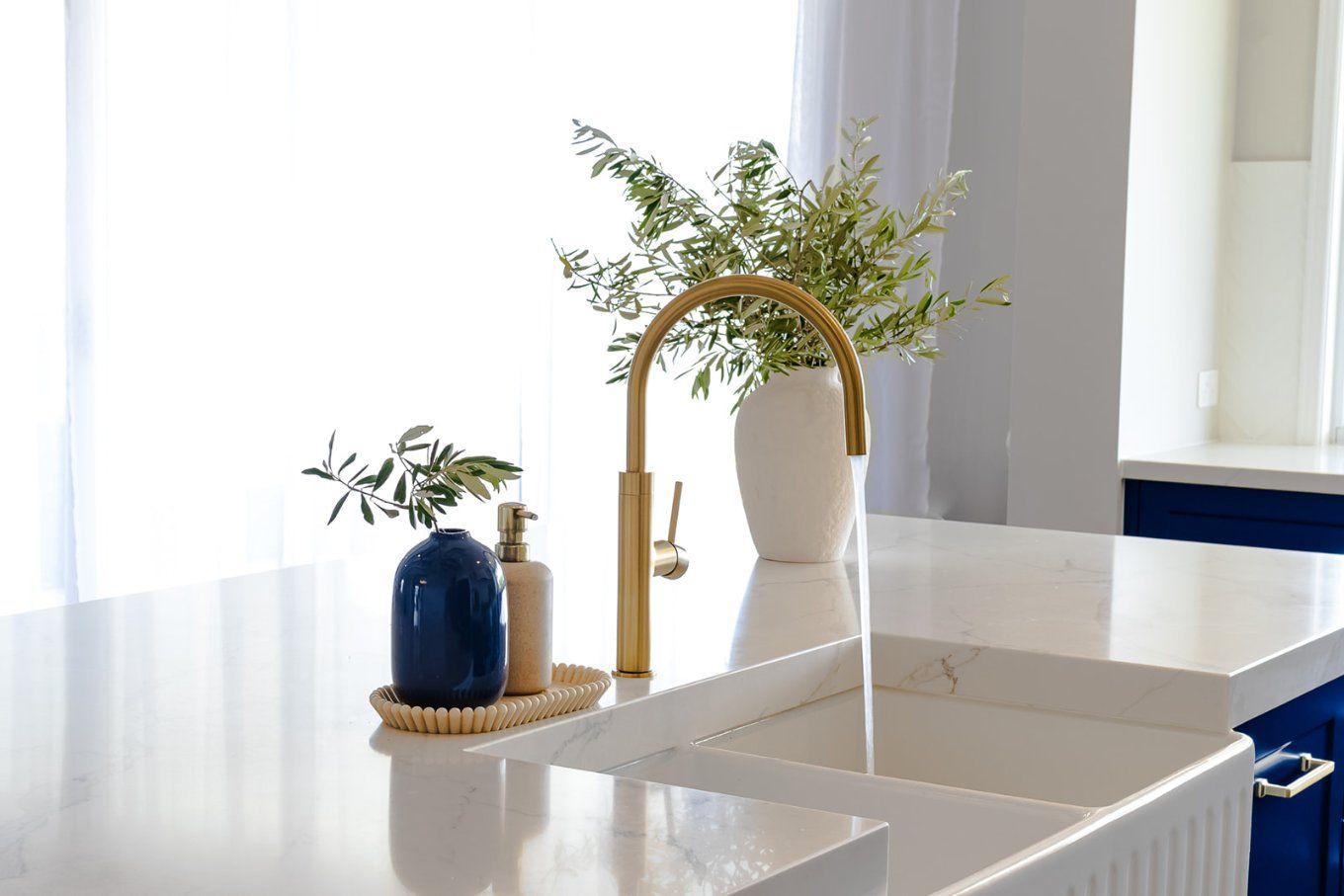 A Gold Faucet Pours Water Into a White Farmhouse Sink on a Bright, White Countertop — Truline Cabinets In Tuchekoi, QLD