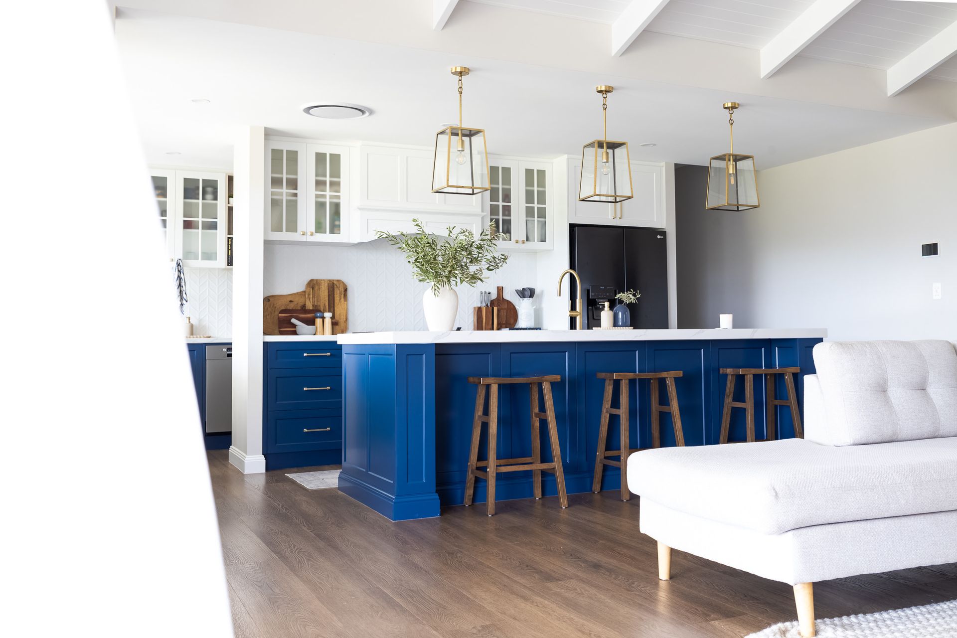 Blue and white kitchen with a large island, gold pendant lights, and wooden stools. A white couch sits in the foreground.