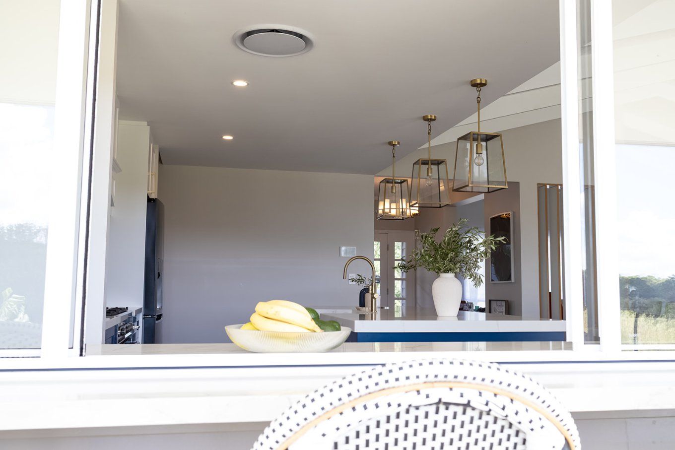 Kitchen Interior Viewed Through a Window, Featuring Fruit on a Counter and Hanging Pendant Lights. the Scene is Bright and Airy — Truline Cabinets In Tuchekoi, QLD