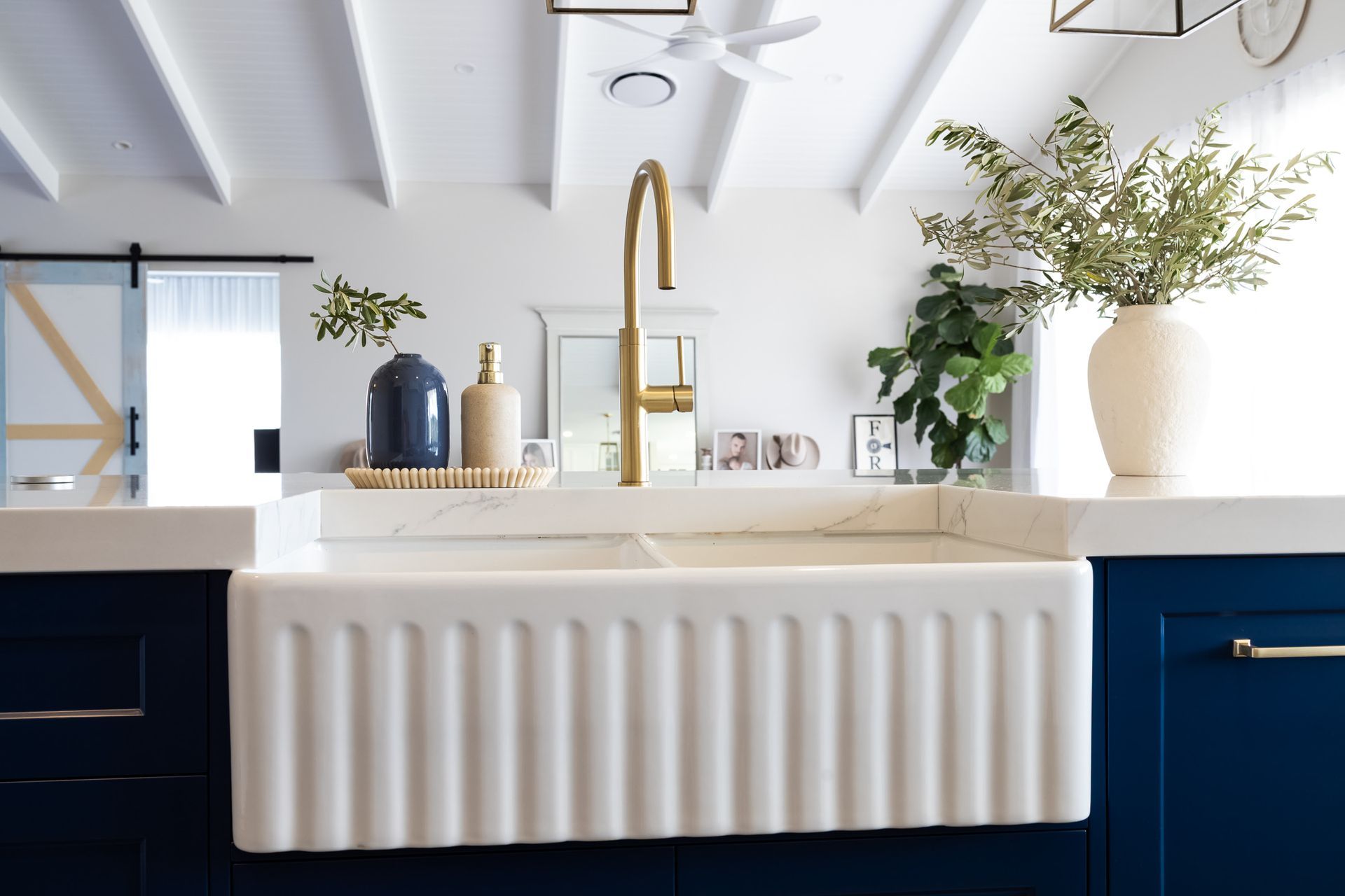 White farmhouse sink with gold faucet, on a navy blue cabinet. A vase with greenery and a blue bottle sit on the countertop.