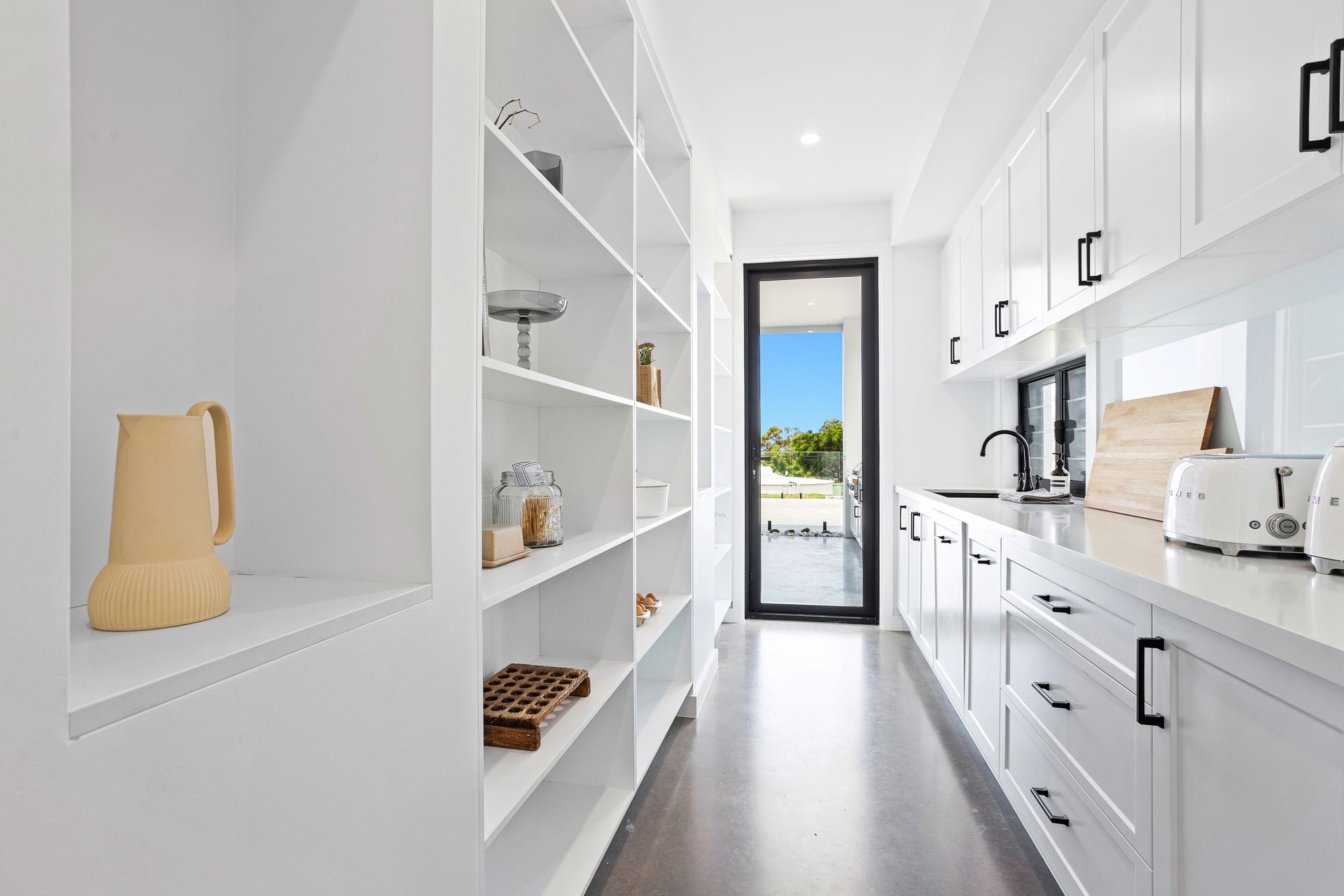 White pantry with shelves on one side and cabinets with a sink on the other, leading to an open door.