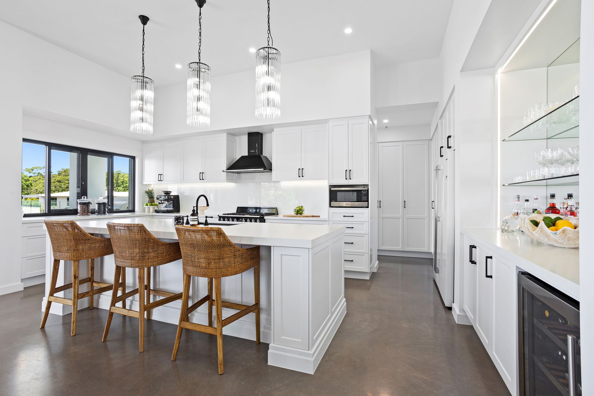 Modern white kitchen with island, wicker bar stools, and pendant lights. Features built-in appliances and a wine fridge.