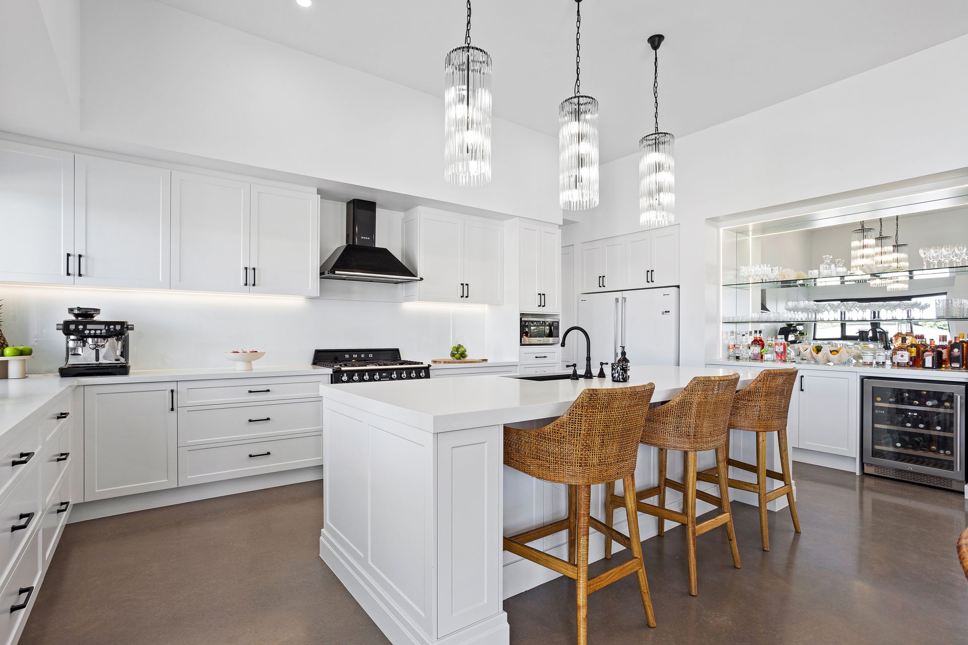 White modern kitchen with island, bar stools, hanging lights, and built-in wine fridge.