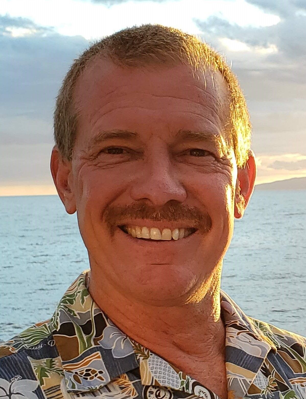 A man with a mustache is smiling in front of the ocean.
