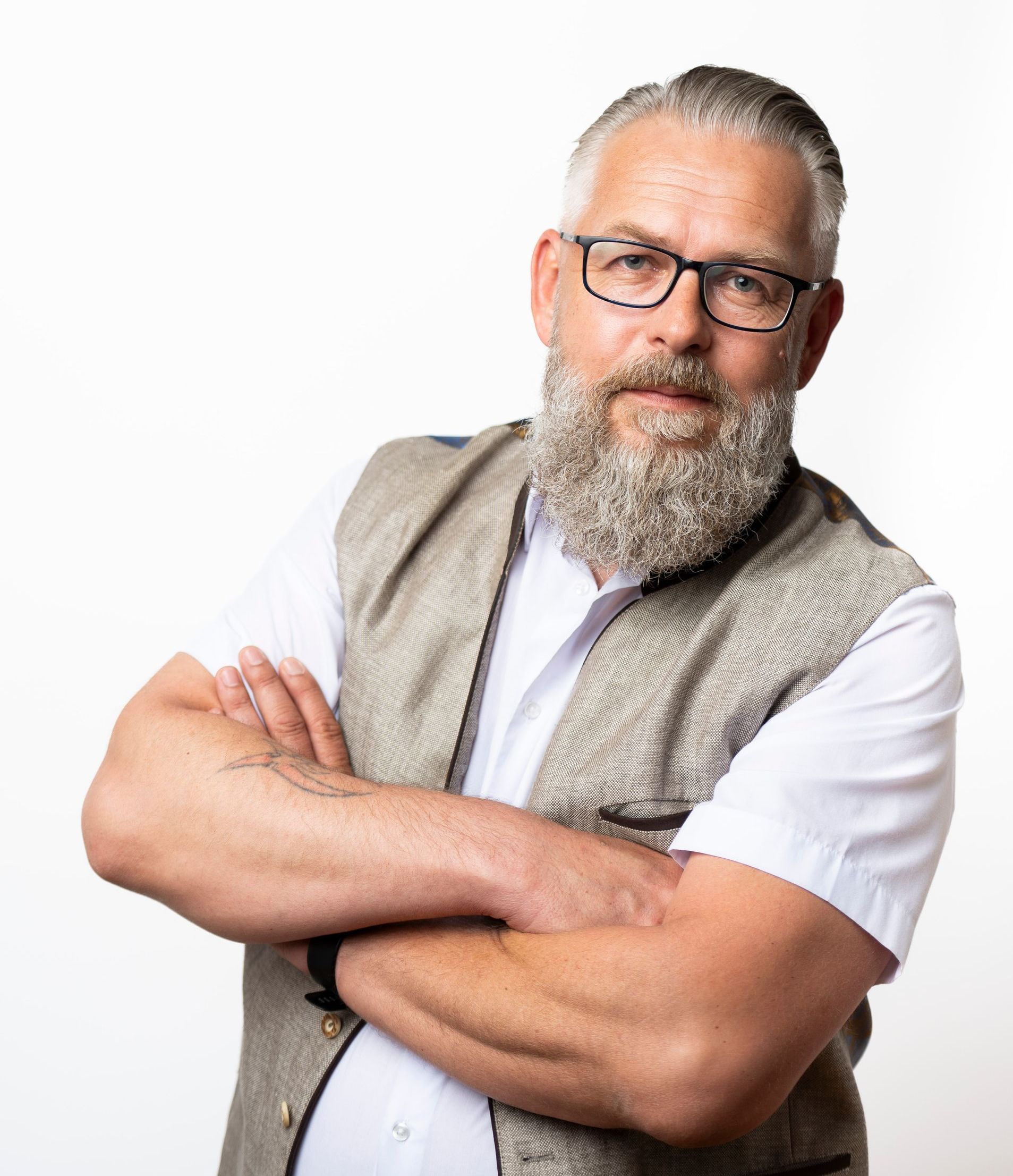 Mann mit grauem Bart und Brille, verschränkten Armen, Weste und weißem Hemd, vor weißem Hintergrund.