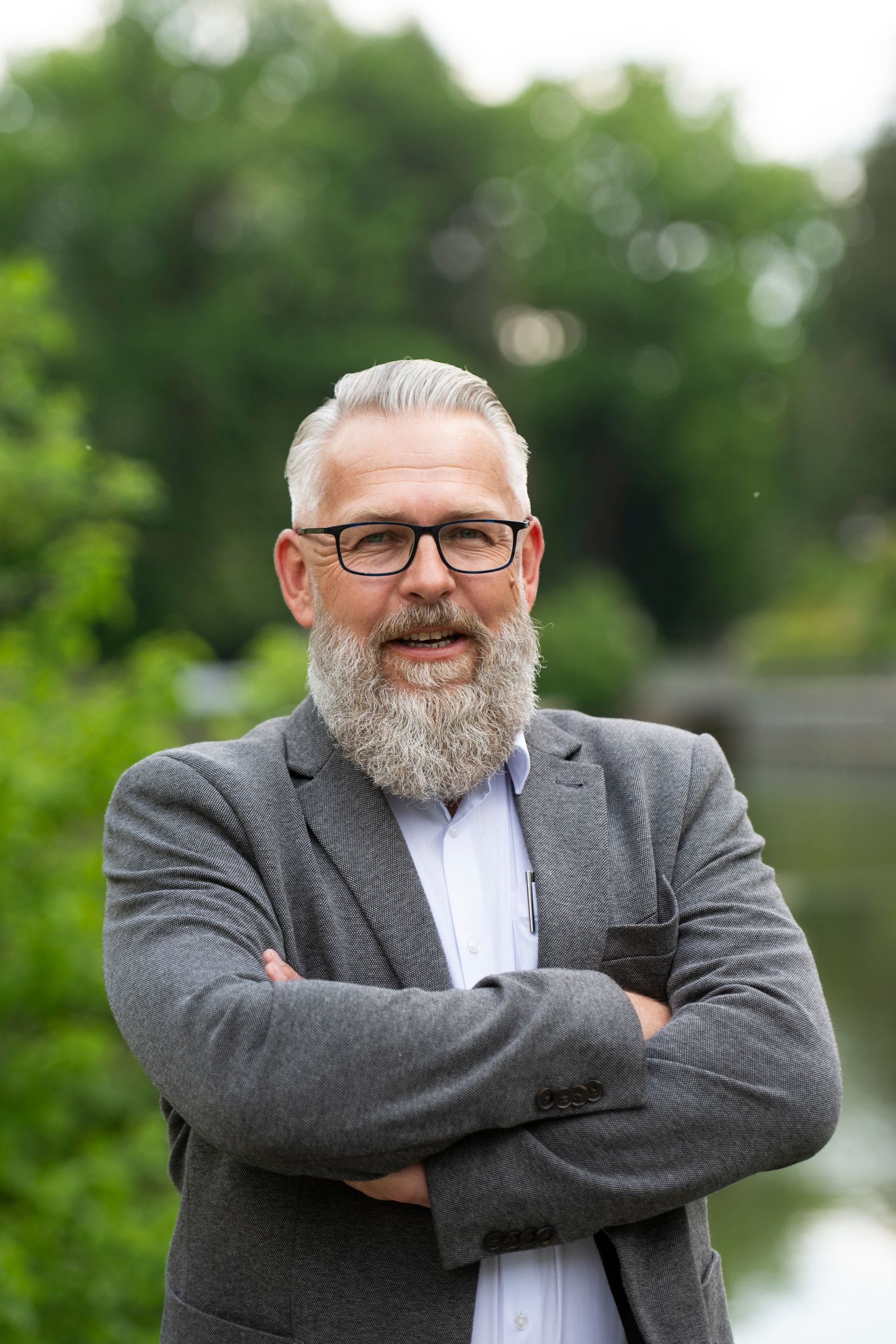 Mann mit grauem Bart, Brille und Blazer lächelt im Freien, die Arme verschränkt.
