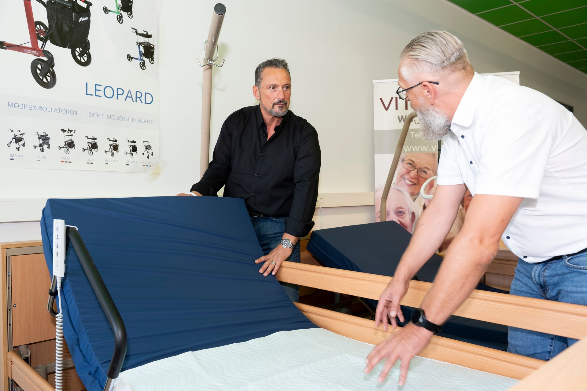 Zwei Männer inspizieren ein verstellbares Krankenhausbett in einem Ausstellungsraum mit Mobilitätshilfen und Produktdisplays.