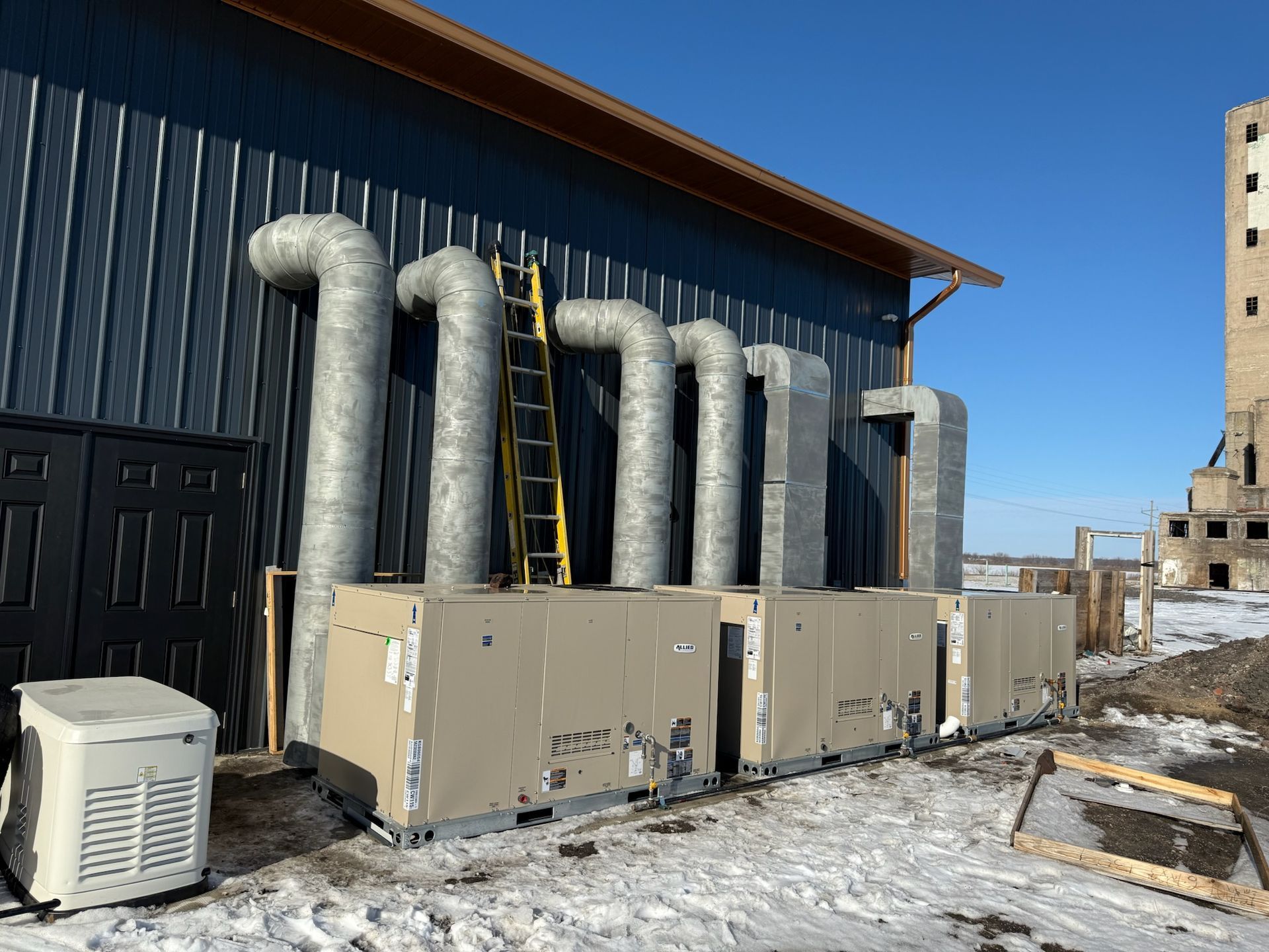 HVAC units on a building exterior with insulated ducts and a ladder. Snow on the ground, bright sunlight.