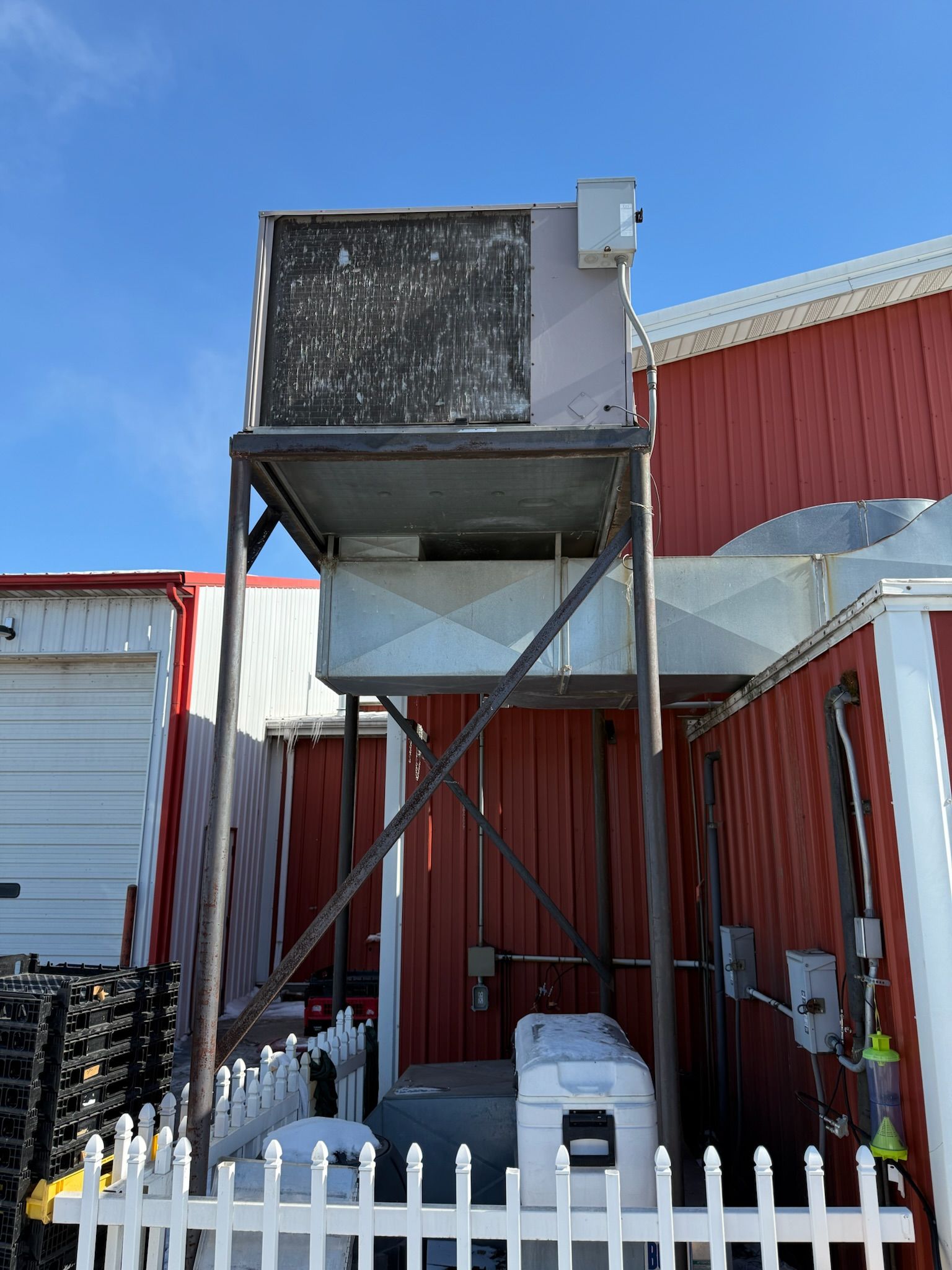 Metal HVAC unit on a tall stand next to a red building with white fence and blue sky.