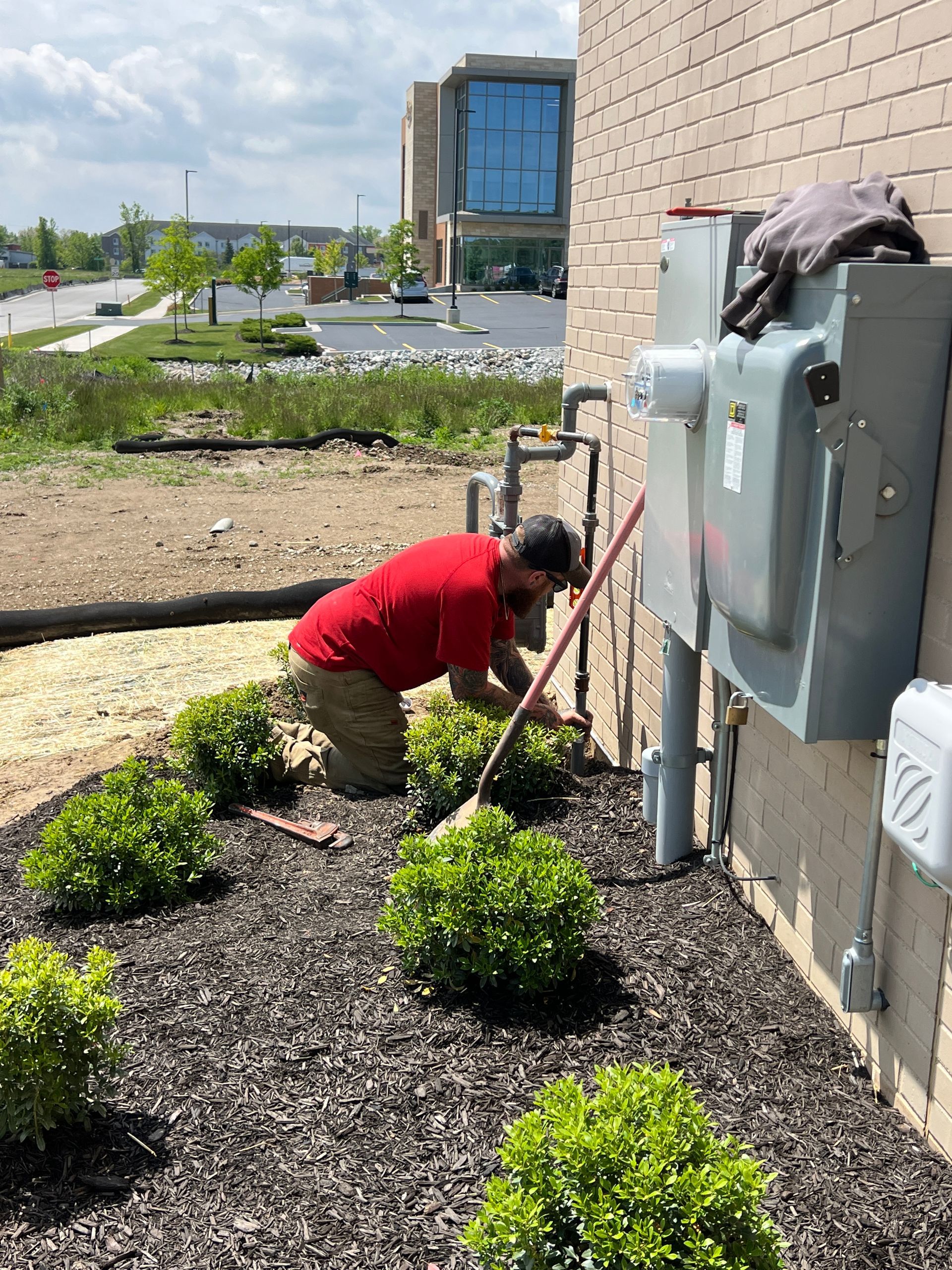 Man in red shirt tending bushes next to a building and utility boxes.