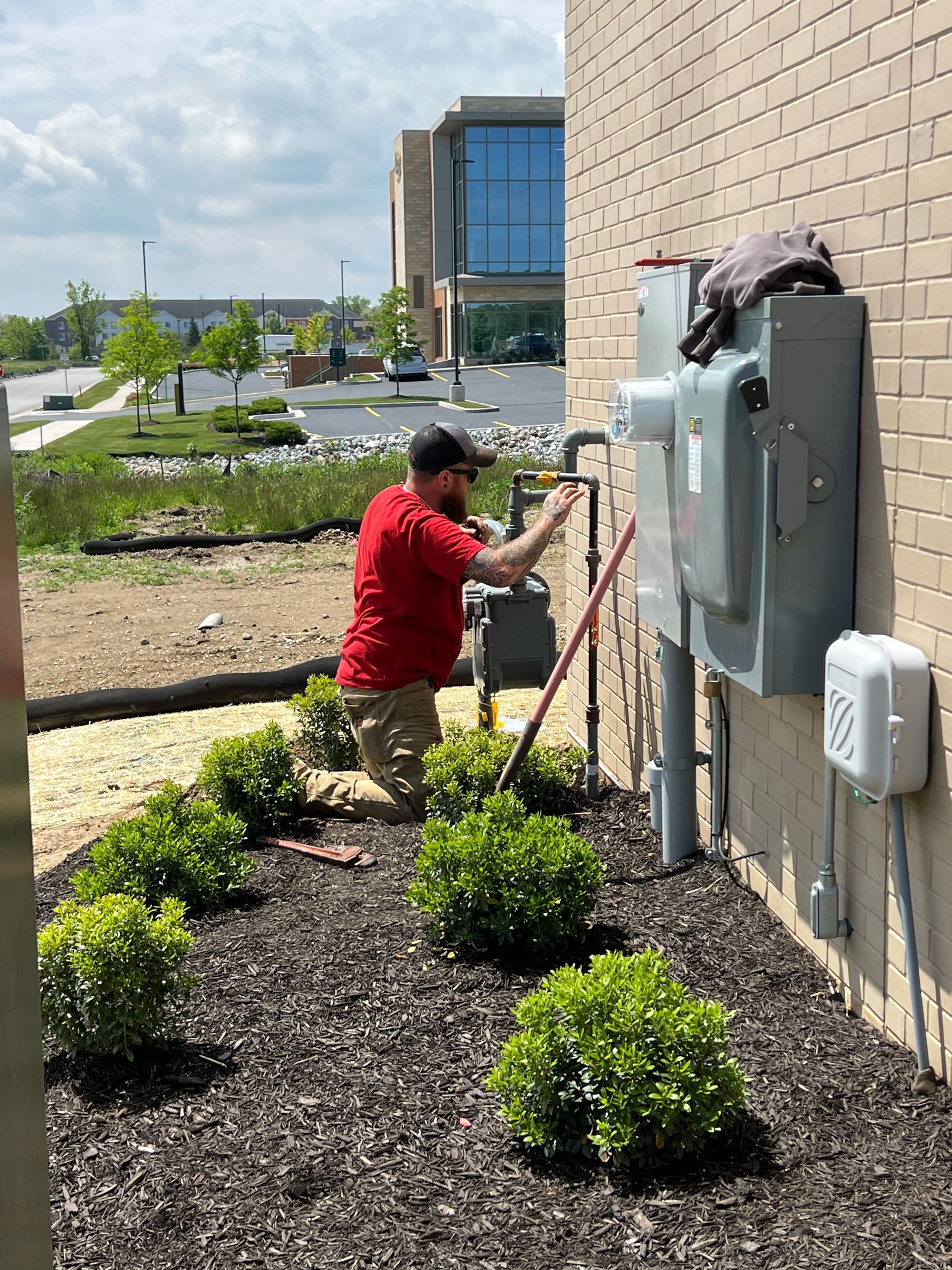 Man in red shirt working on gas meter on a building's exterior wall, in front of a landscaped area.