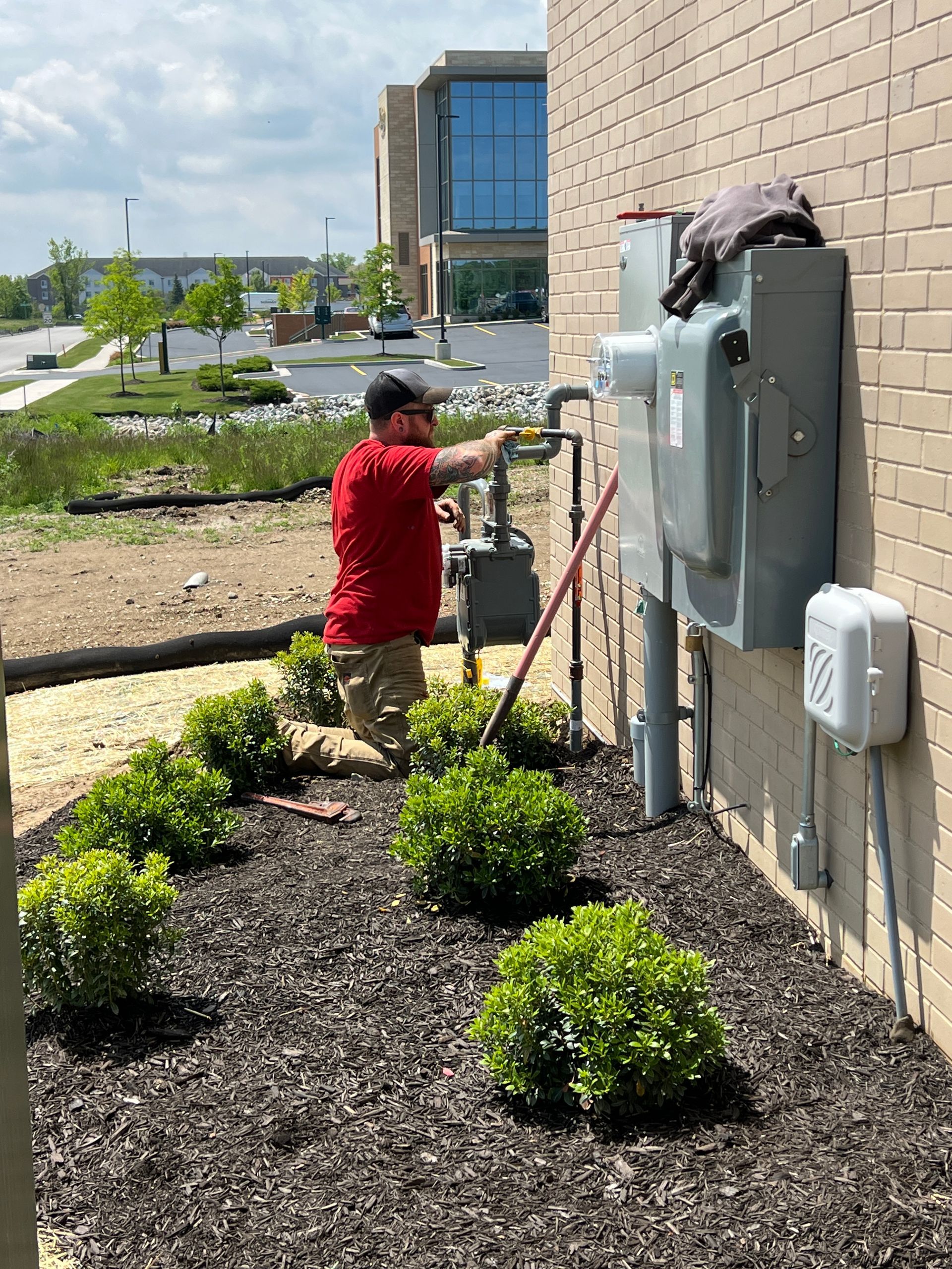 A person in a red shirt adjusts a gas meter on a building's brick exterior; garden and street visible.