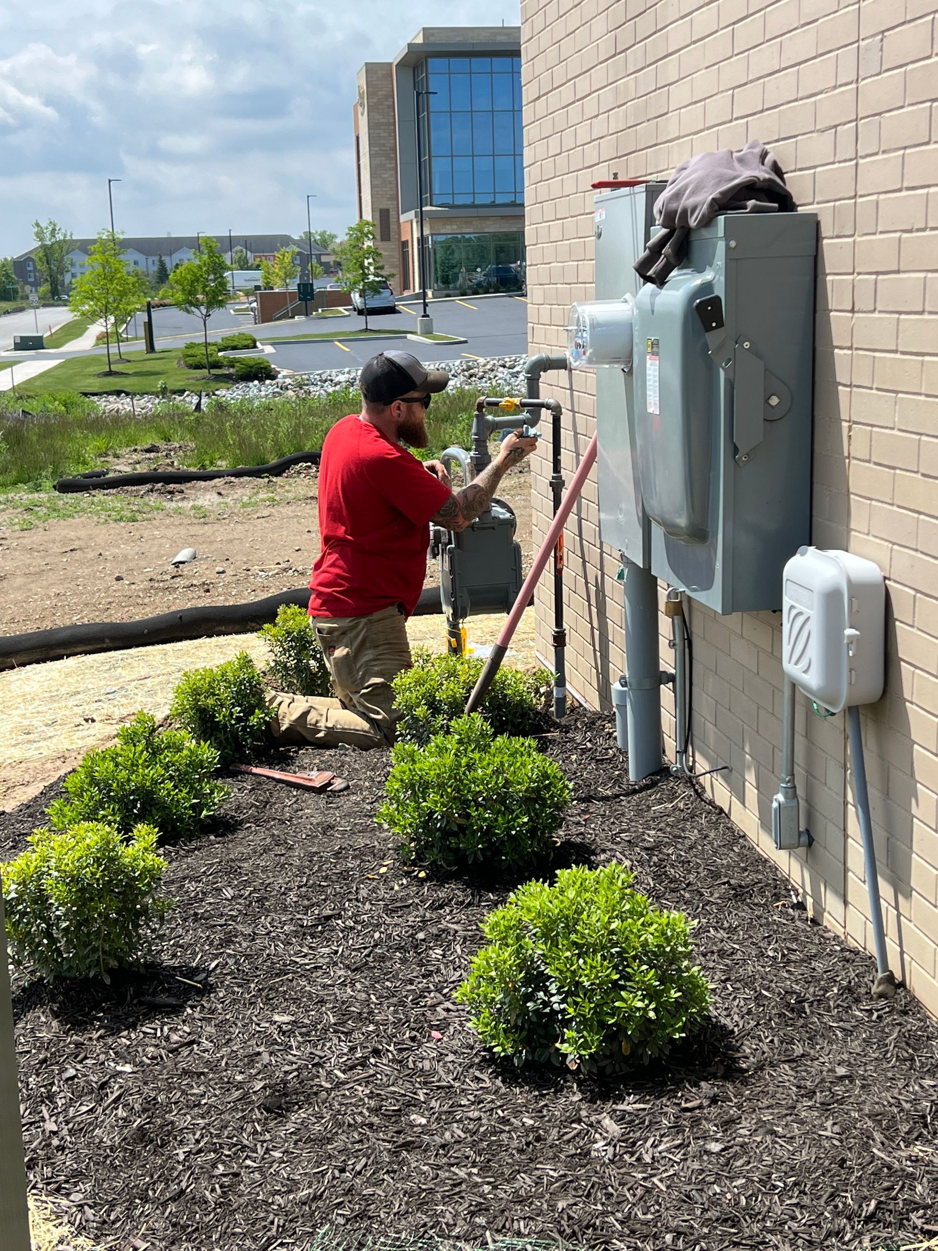 A person in a red shirt works on a gas meter box attached to a brick building.