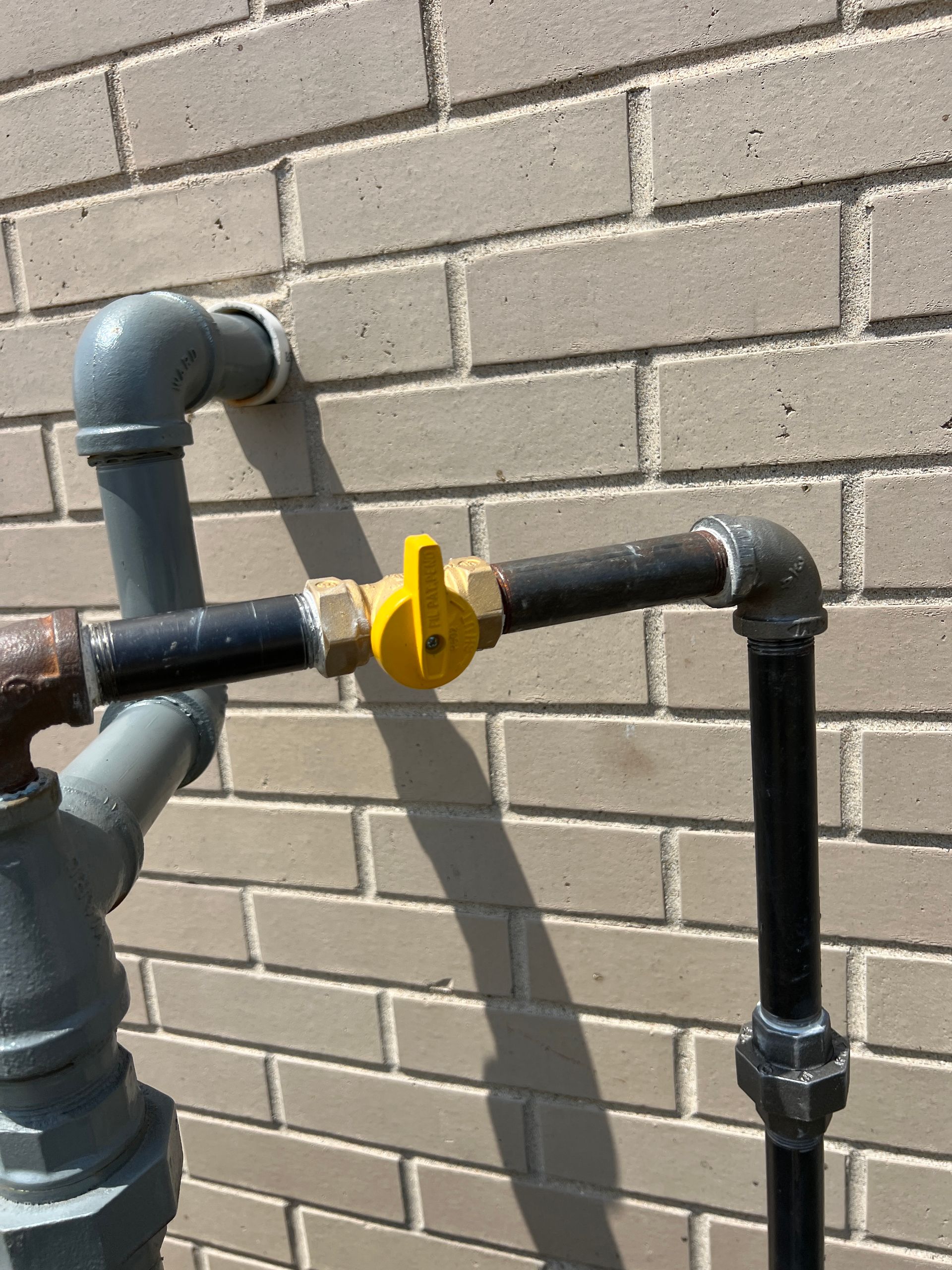 Gas shut-off valve on brick wall. Yellow handle on black pipe with gray and black elbows.