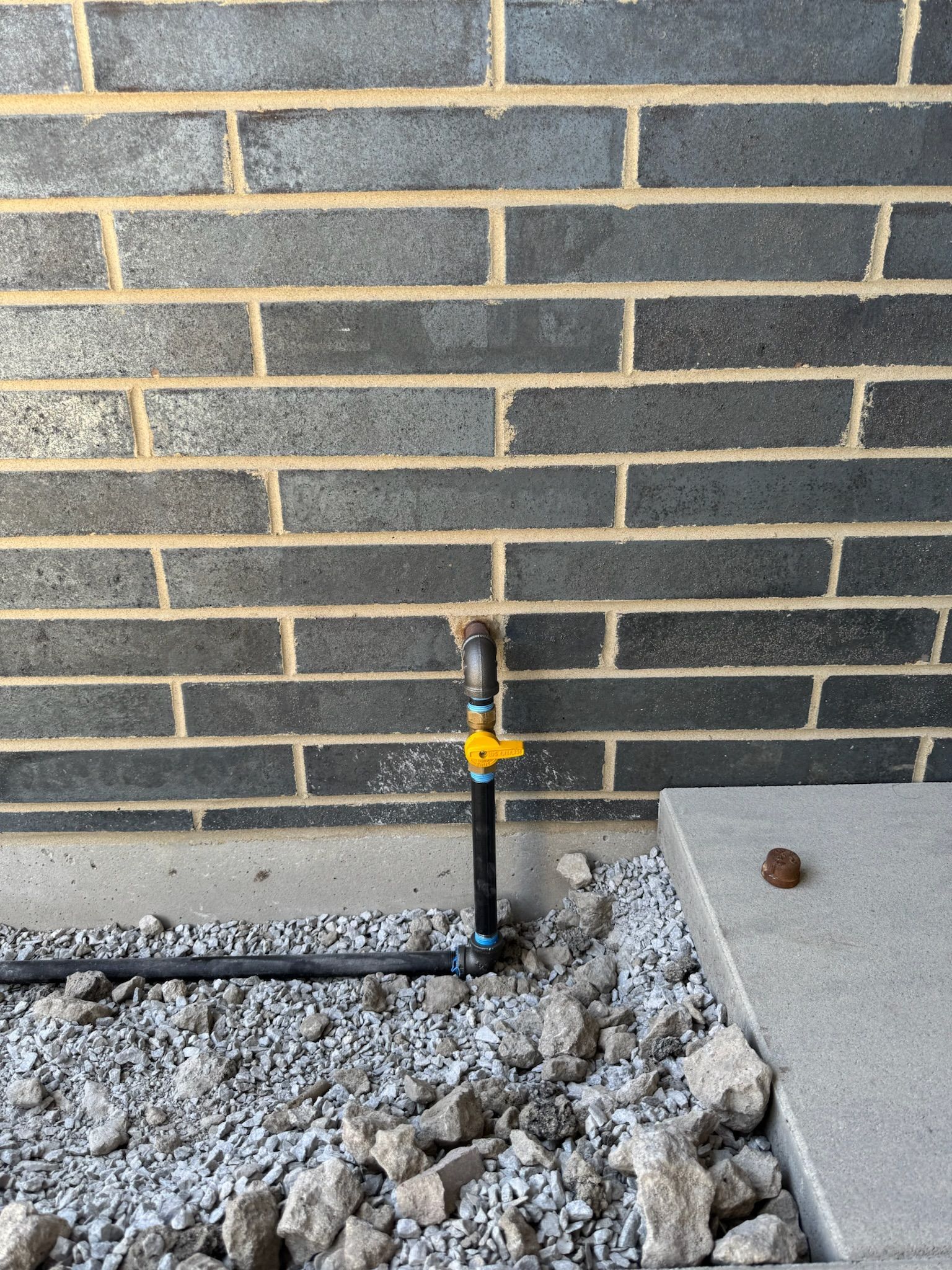 Black gas pipe with yellow handle, gravel, and brick wall.