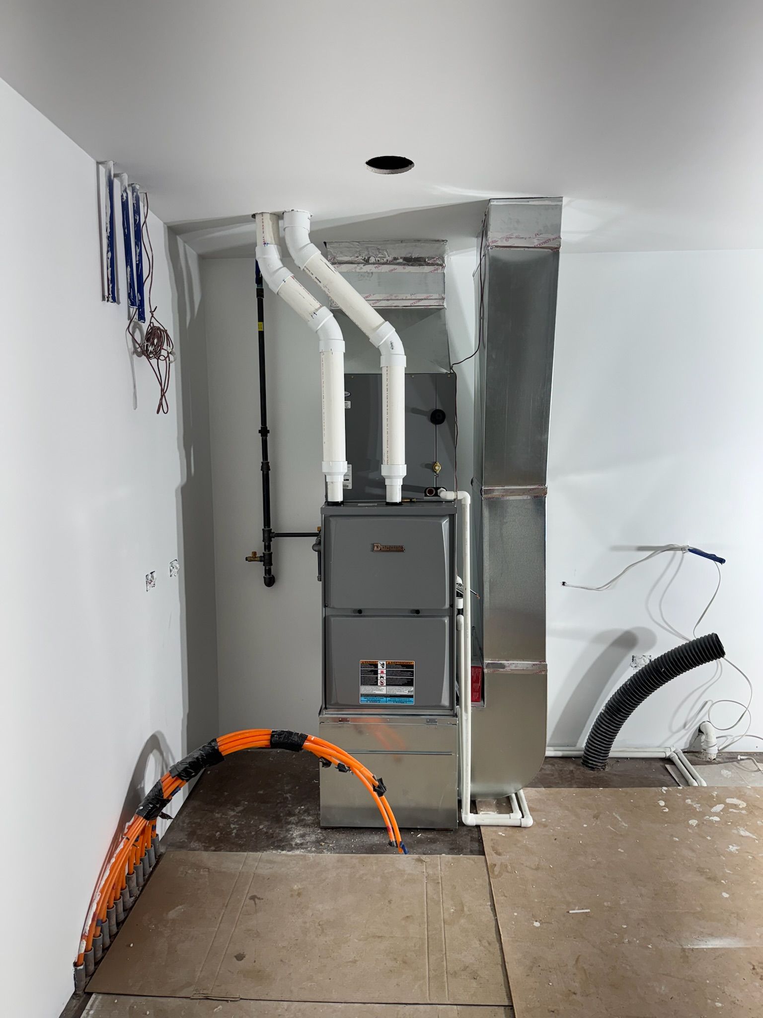 Furnace in a utility room. White and gray pipes, metallic ductwork, and electrical wiring are visible.