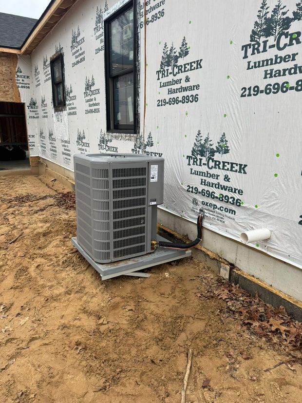 HVAC unit installed outdoors against a partially built house, on a metal base, in a dirt yard.