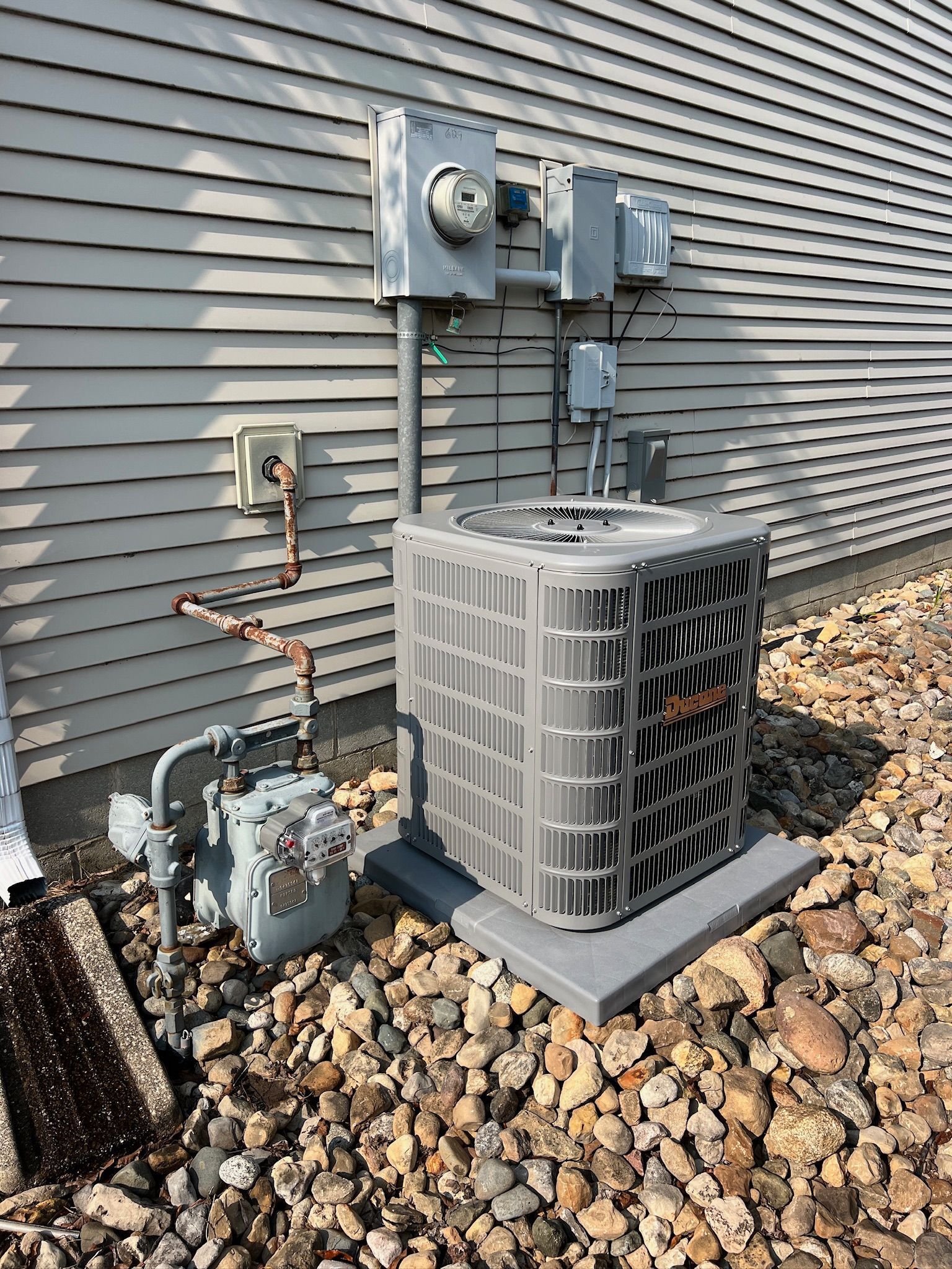 Outdoor HVAC unit and electrical components next to a house with gray siding, set on gravel.