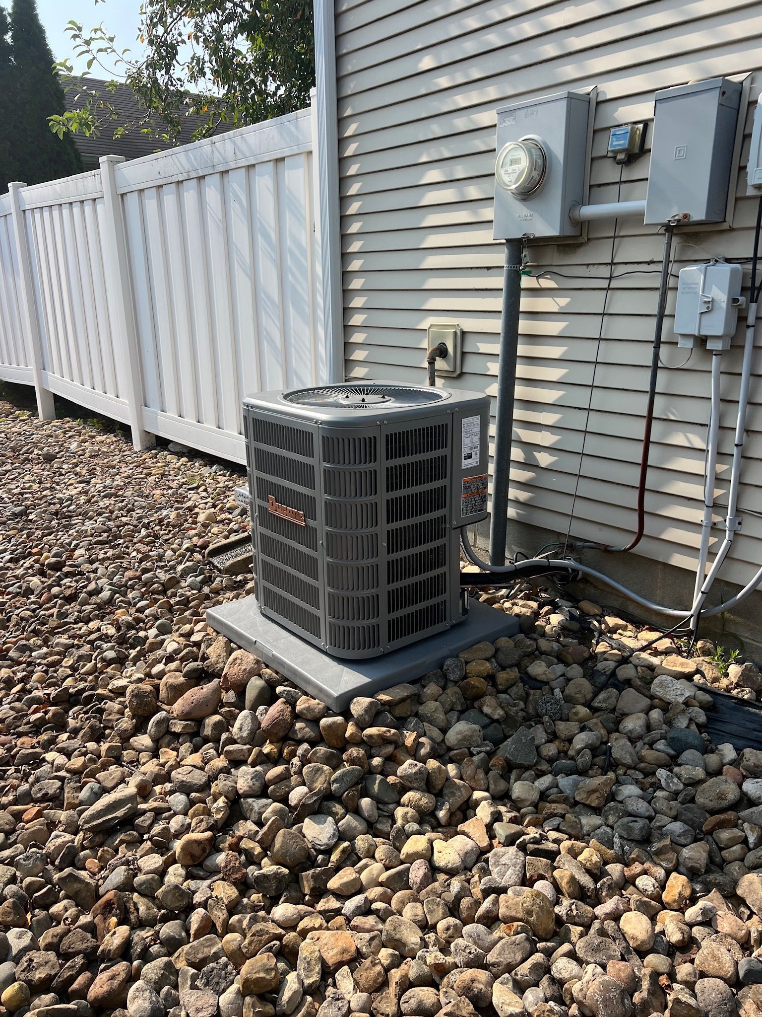 Air conditioning unit next to a house with electrical boxes and a white fence, surrounded by gravel.