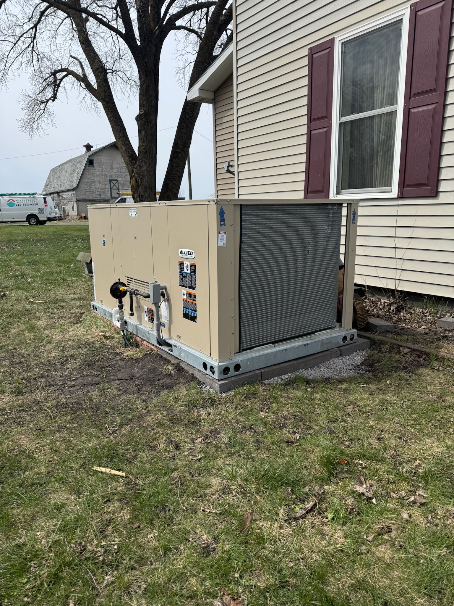 Tan generator unit next to a house with maroon shutters and a leafless tree.
