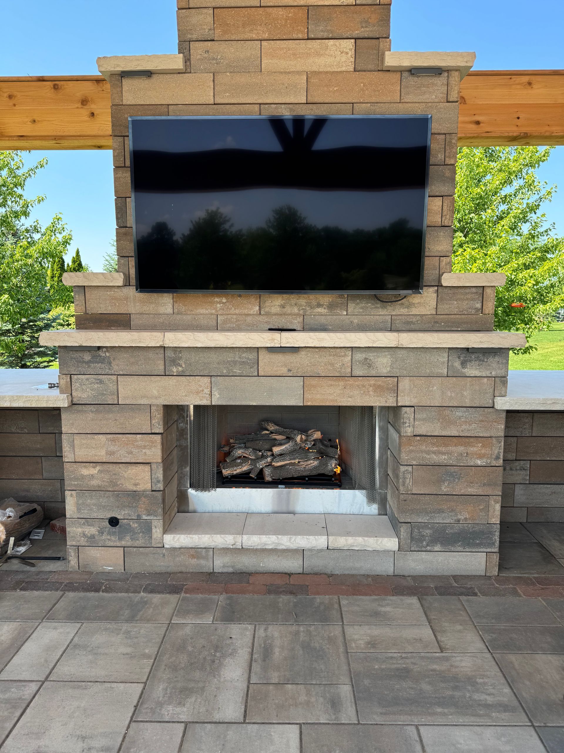 Outdoor fireplace with mounted TV, made of stone. Includes wood and flames, set on a patio with a blue sky background.