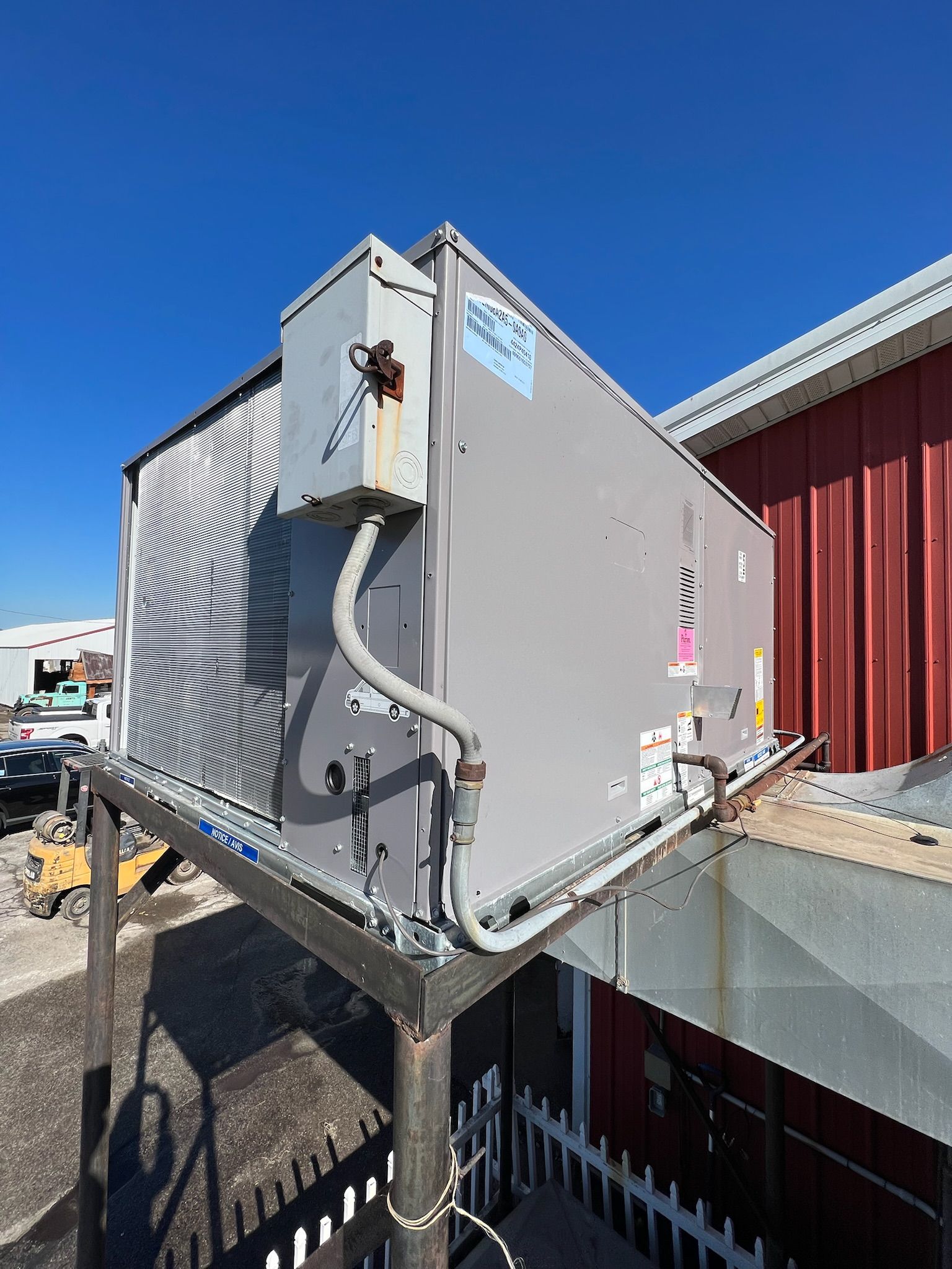 Gray HVAC unit on a metal frame, mounted on a rooftop with a red building and blue sky.