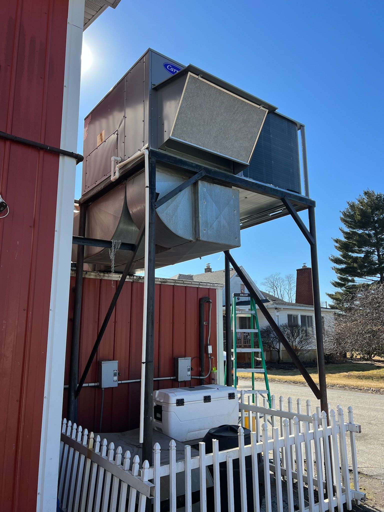 Metal HVAC unit on a support structure outside a red building with a white picket fence.
