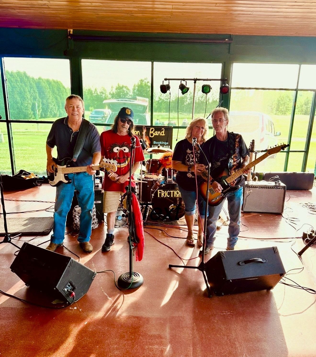 Band performing outside on a stage. Four people with instruments, a drum set in the background, and a green landscape.